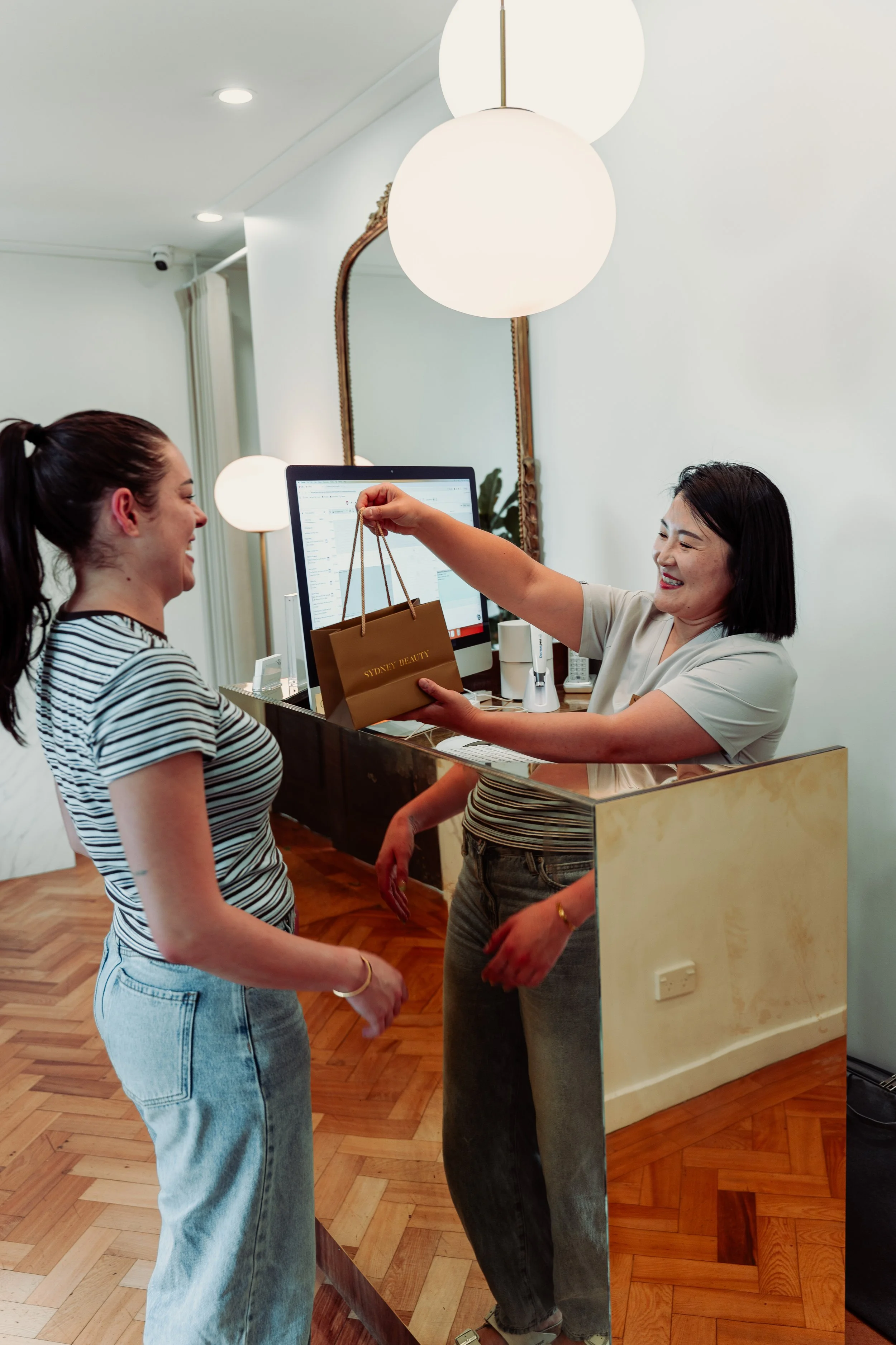 A woman at a reception desk handing a shopping bag to another woman. Both are smiling indoors with wooden flooring and modern lighting.