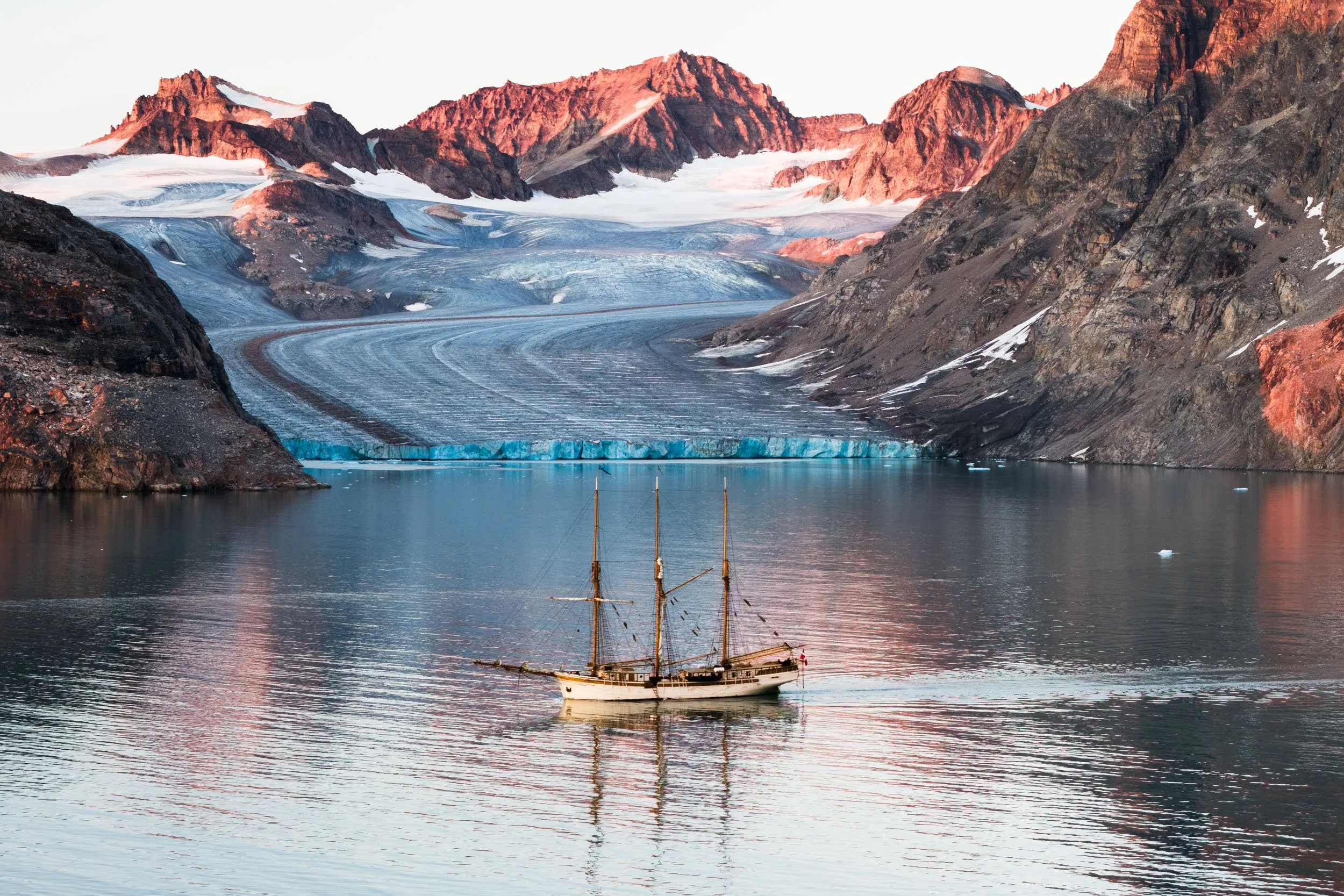 S/V Linden on calm water with glacier-covered mountains and a glacier in the background, illuminated by soft sunset light.