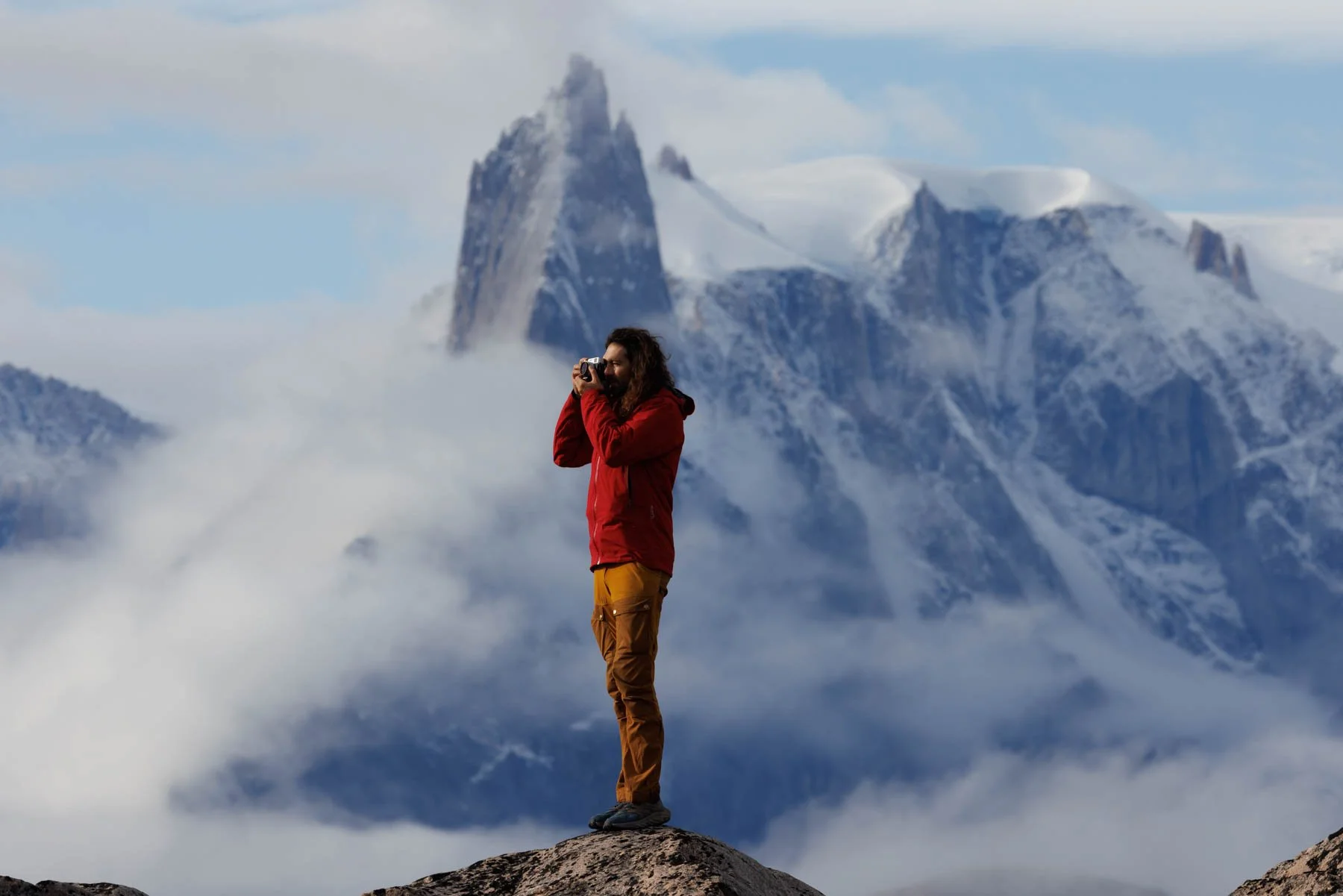 Photographer Karim Iliya  long hair and a beard wearing a red jacket and brown pants standing on a rock, taking a photograph with a camera, with snow-capped mountains and clouds in the background.