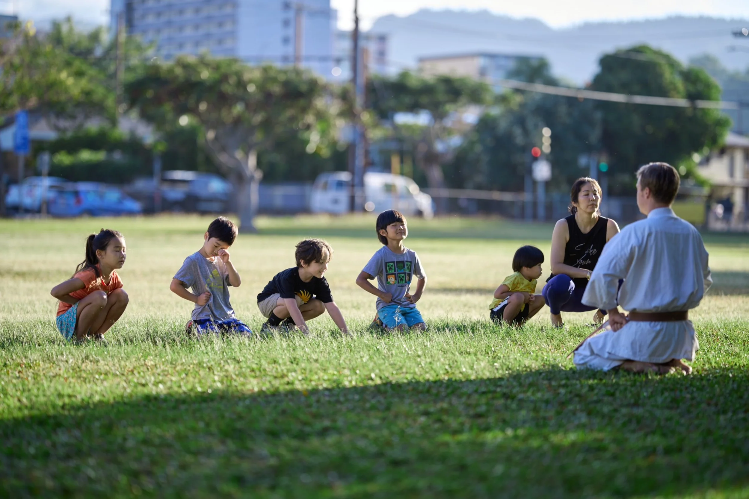 Children participating in a martial arts class outdoors, sitting in a line on the grass as an instructor demonstrates a move.