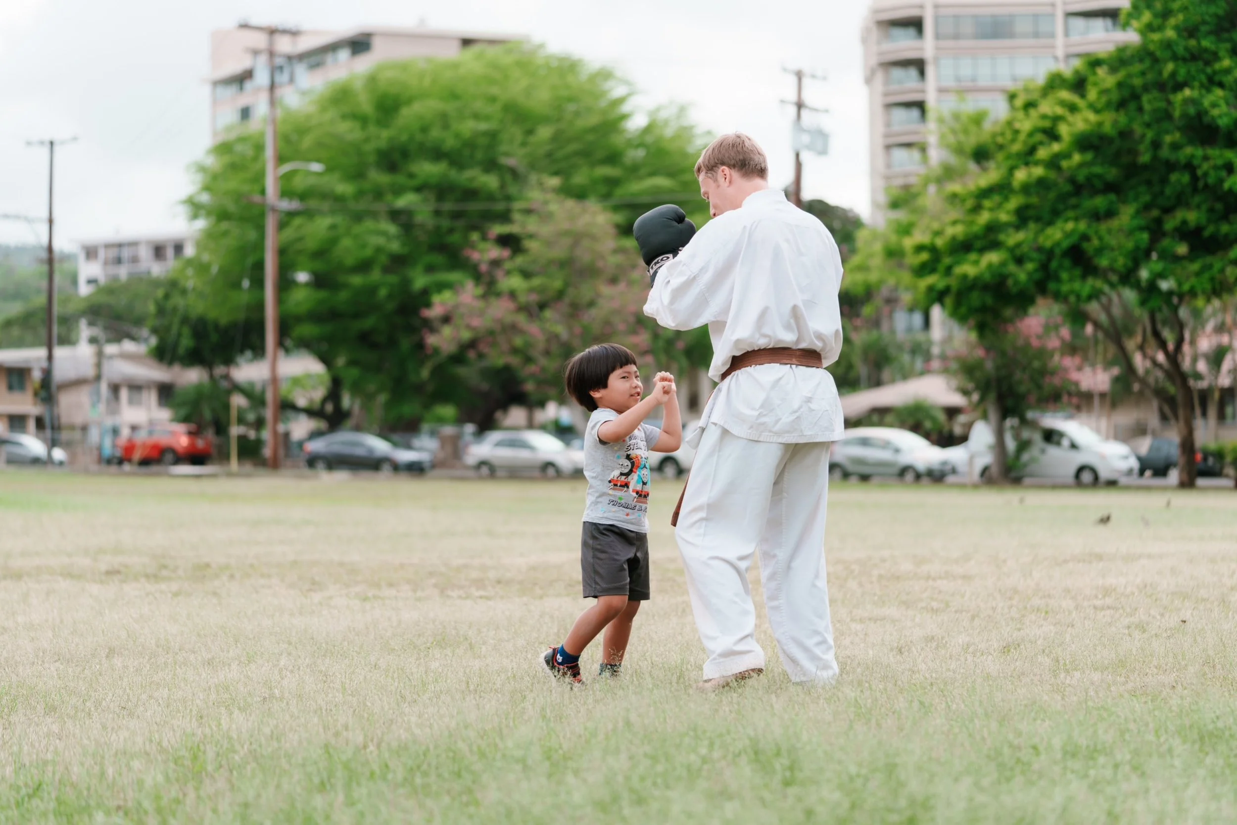 A young boy practicing martial arts with an adult in a park, surrounded by trees and residential buildings.
