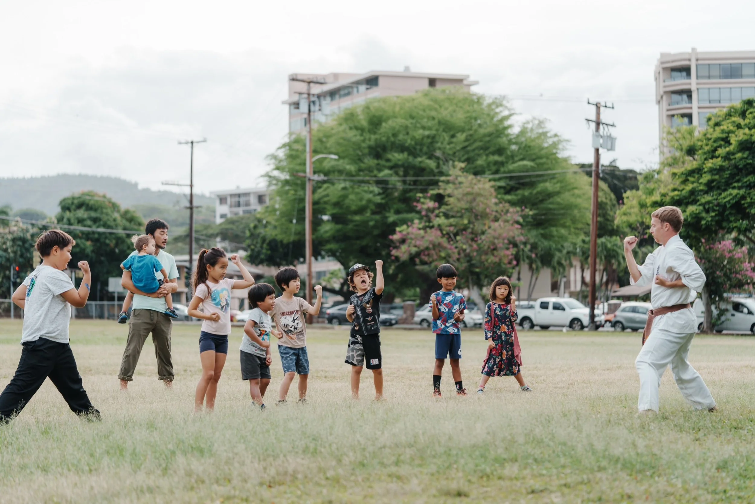 Kids and adults practicing martial arts outdoors in a park, with trees and buildings in the background.