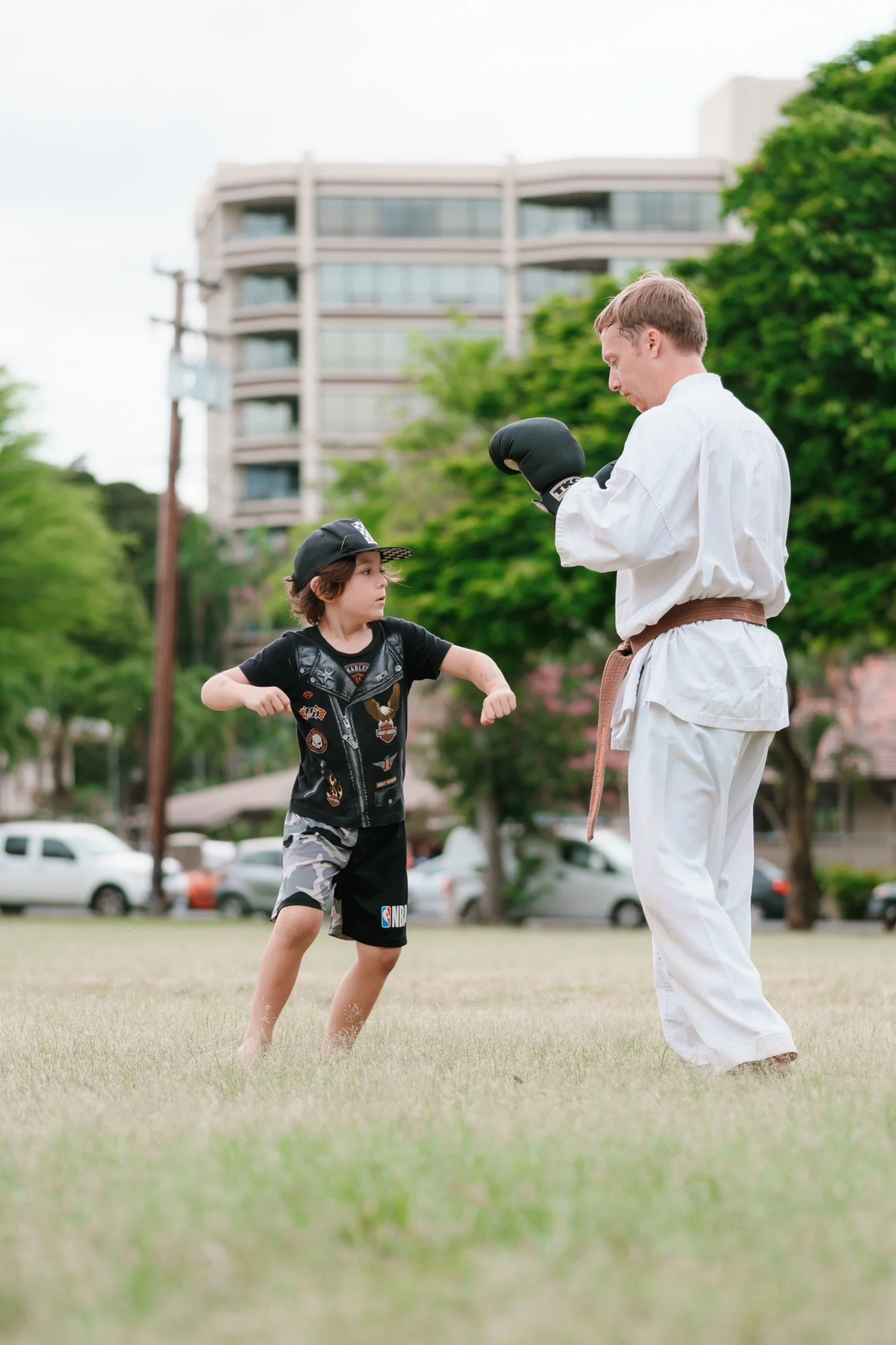 A young boy practicing martial arts with an instructor outdoors in a park, with trees and a building in the background.
