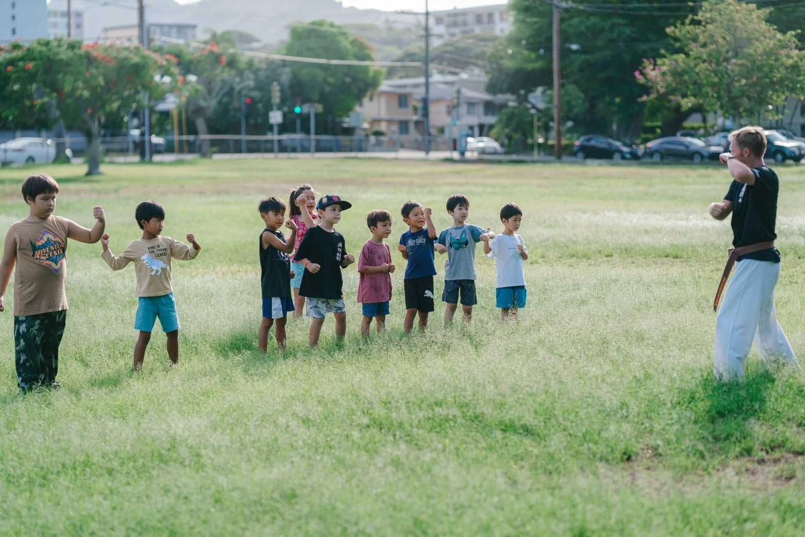 Children participating in a martial arts class outdoors, standing in a line facing an instructor in a park.