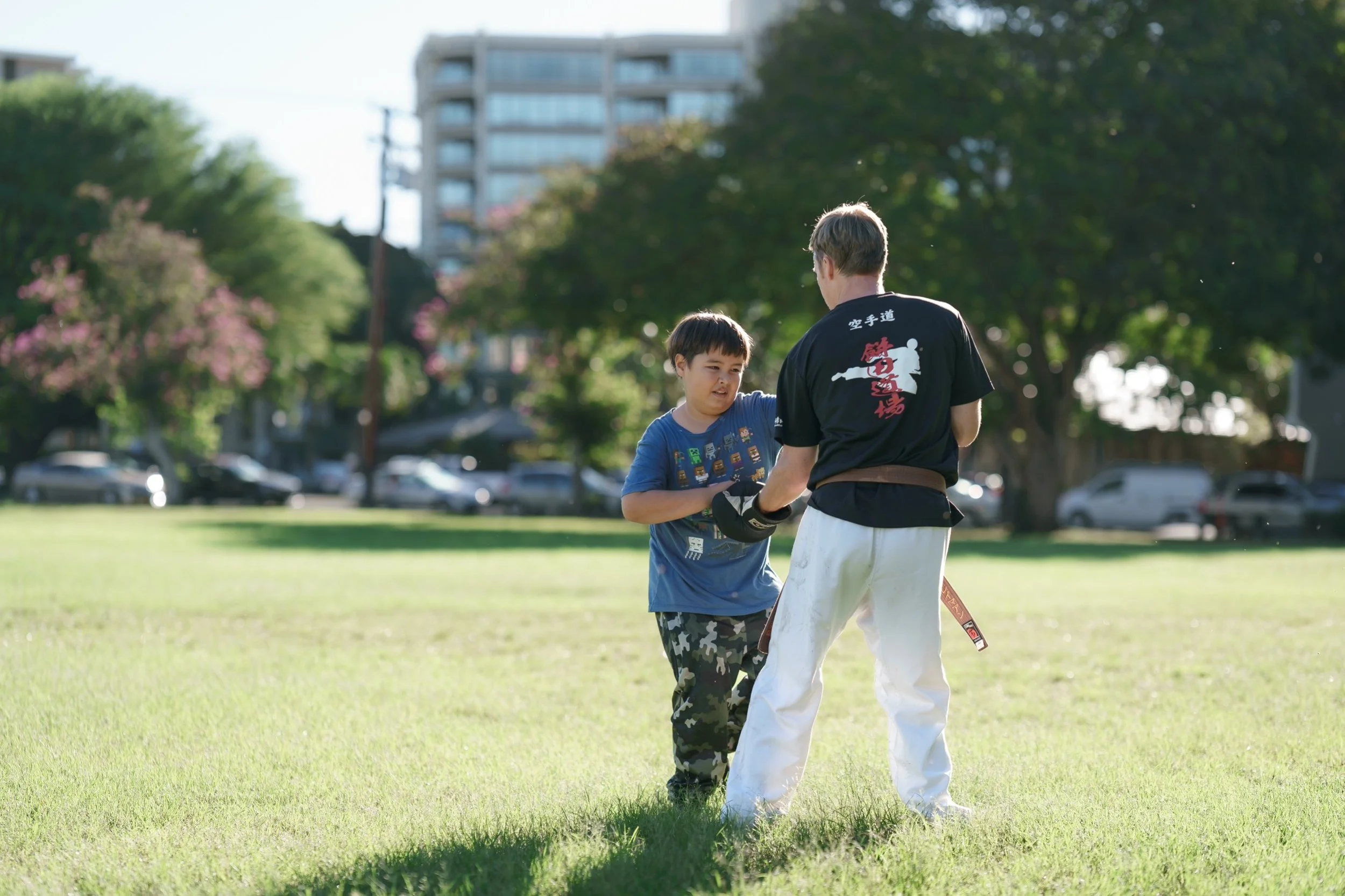 A young boy practicing martial arts with an instructor in a park on a sunny day, with trees and apartment buildings in the background.