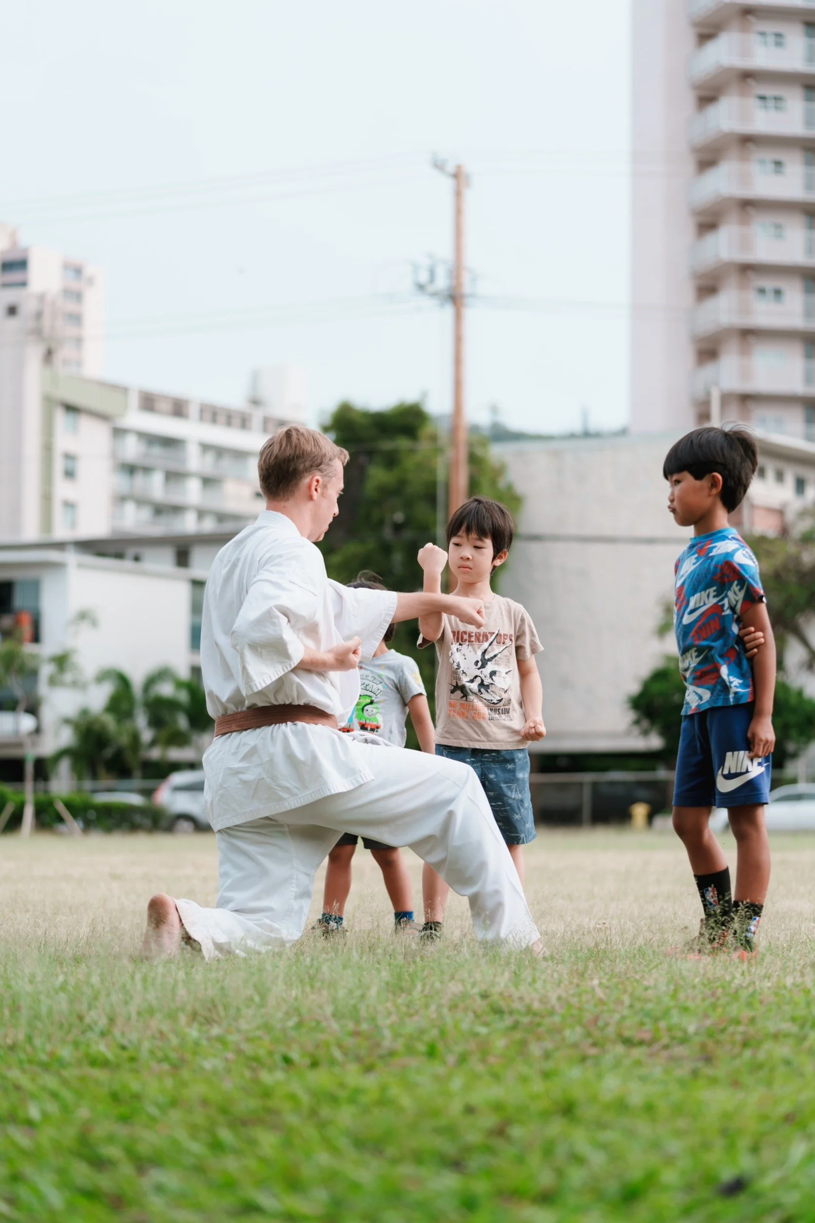 A martial arts instructor demonstrating a technique with children outdoors on a grassy field in an urban area.