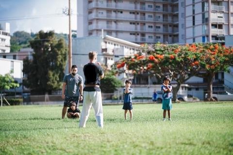 A group of five people, including children and adults, stand in a grassy field in an urban area with buildings and trees in the background. One adult appears to be instructing or talking to the children.