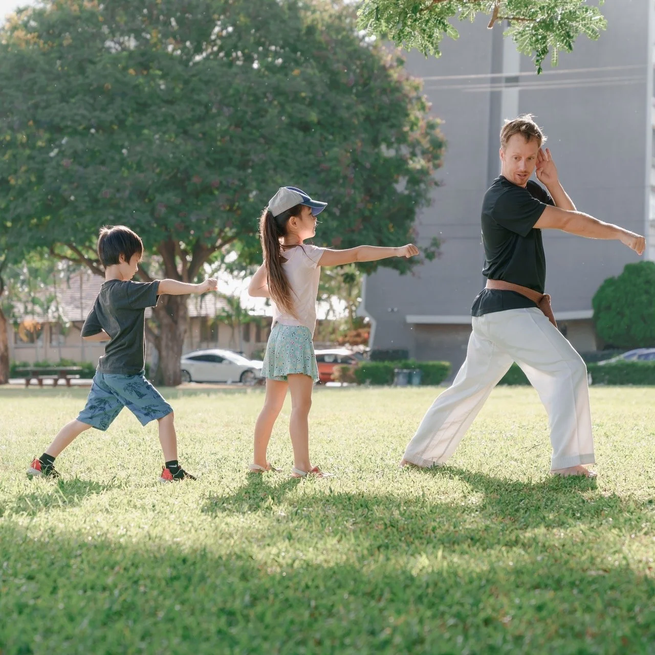 A man and two children practicing martial arts outdoors in a park with trees, grass, and buildings in the background.