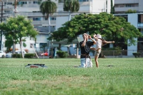 A person kneeling on the grass taking a photo of a woman who is dancing or jumping on a grassy field in a park, with apartment buildings and trees in the background.