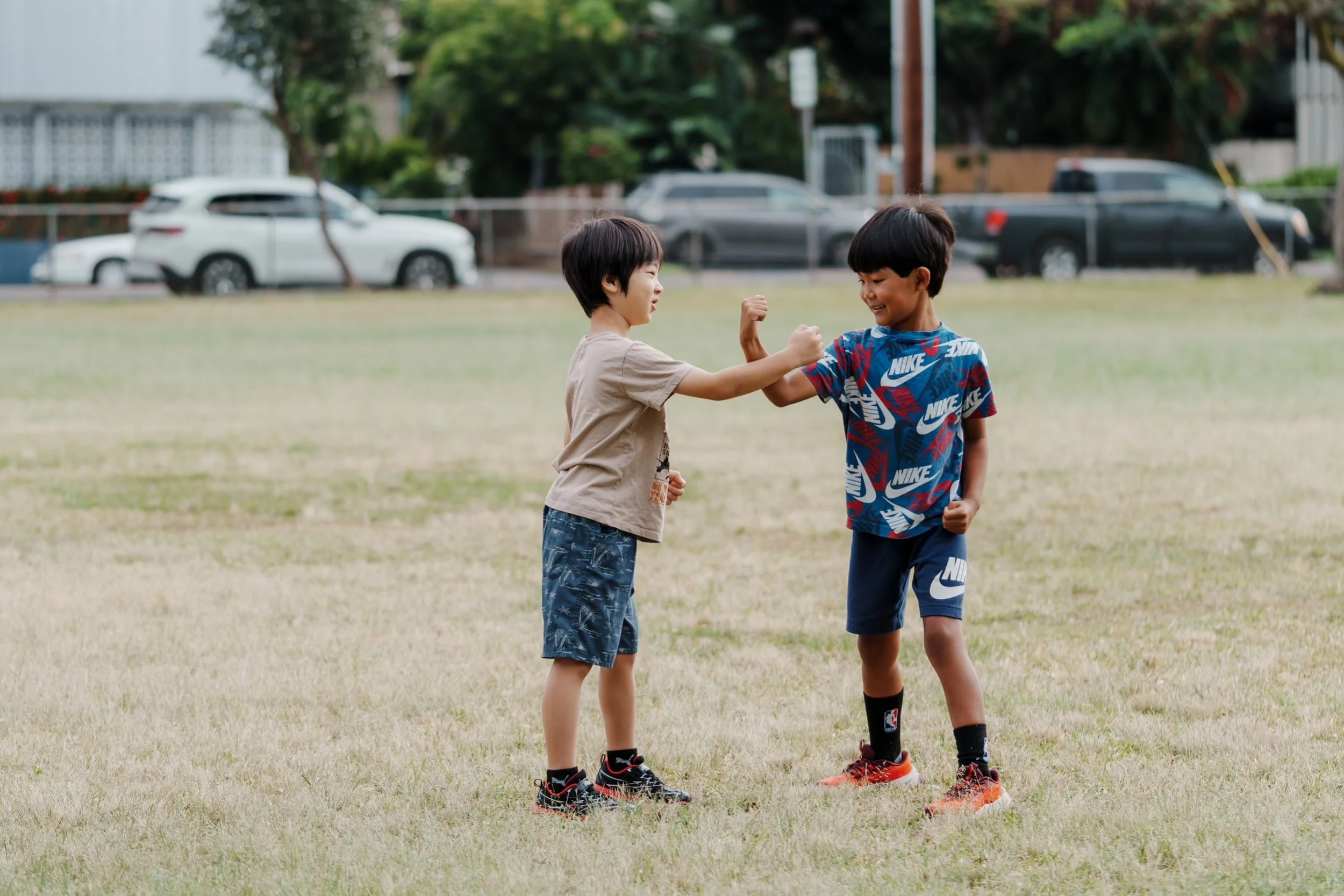 Two boys playing and smiling on a grassy field, bumping fists in a friendly embrace with cars and trees in the background.