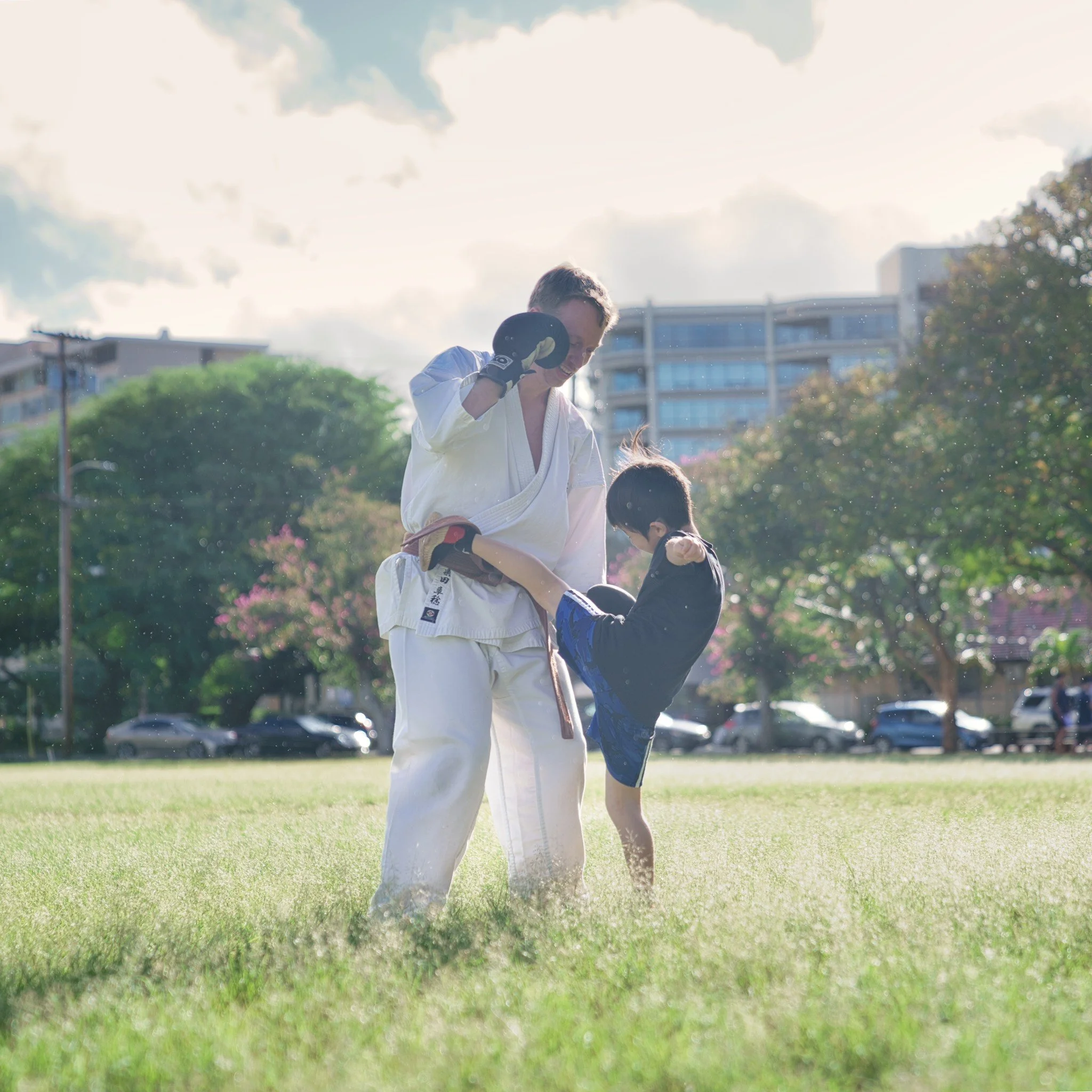 An adult wearing a white karate gi and black gloves practicing martial arts with a young boy in a black and blue gi outdoors on a grassy field with trees and buildings in the background.