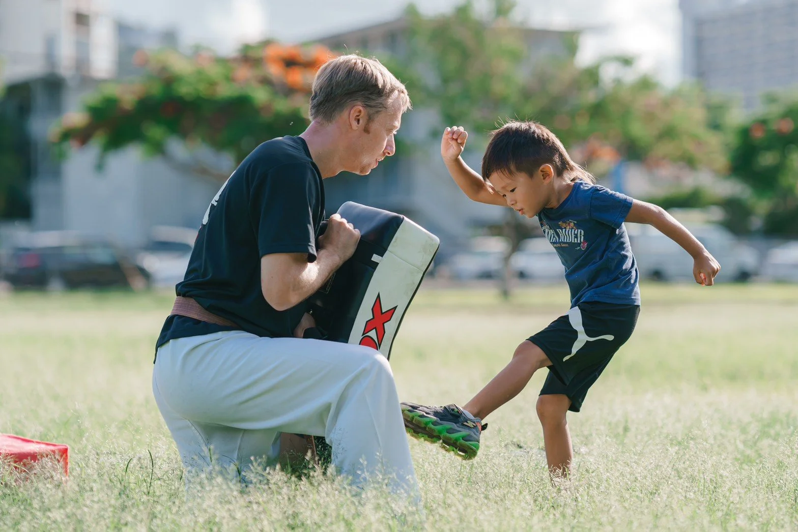 A young boy practicing martial arts with an instructor outdoors in a grassy park, with trees and buildings in the background.