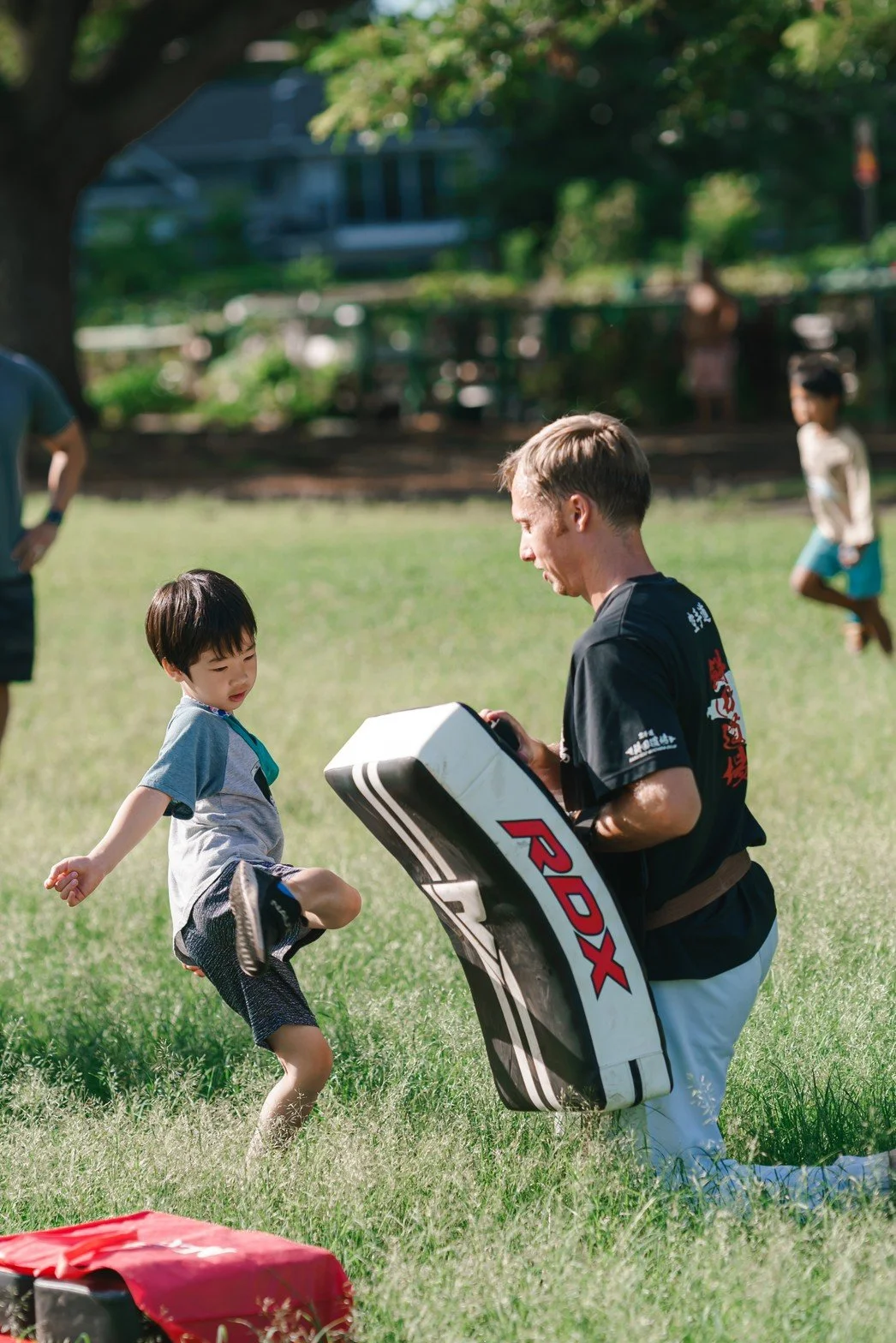A young boy practicing martial arts with an instructor holding a large kicking pad in a park on a sunny day.