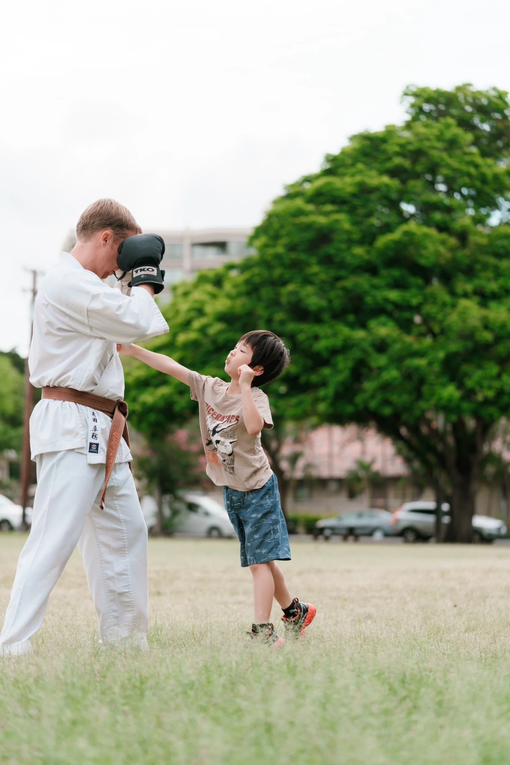 A young boy practicing martial arts with an adult in a park with trees and cars in the background.