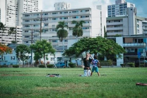 A woman and a child walking on a grassy park with trees and urban buildings in the background.