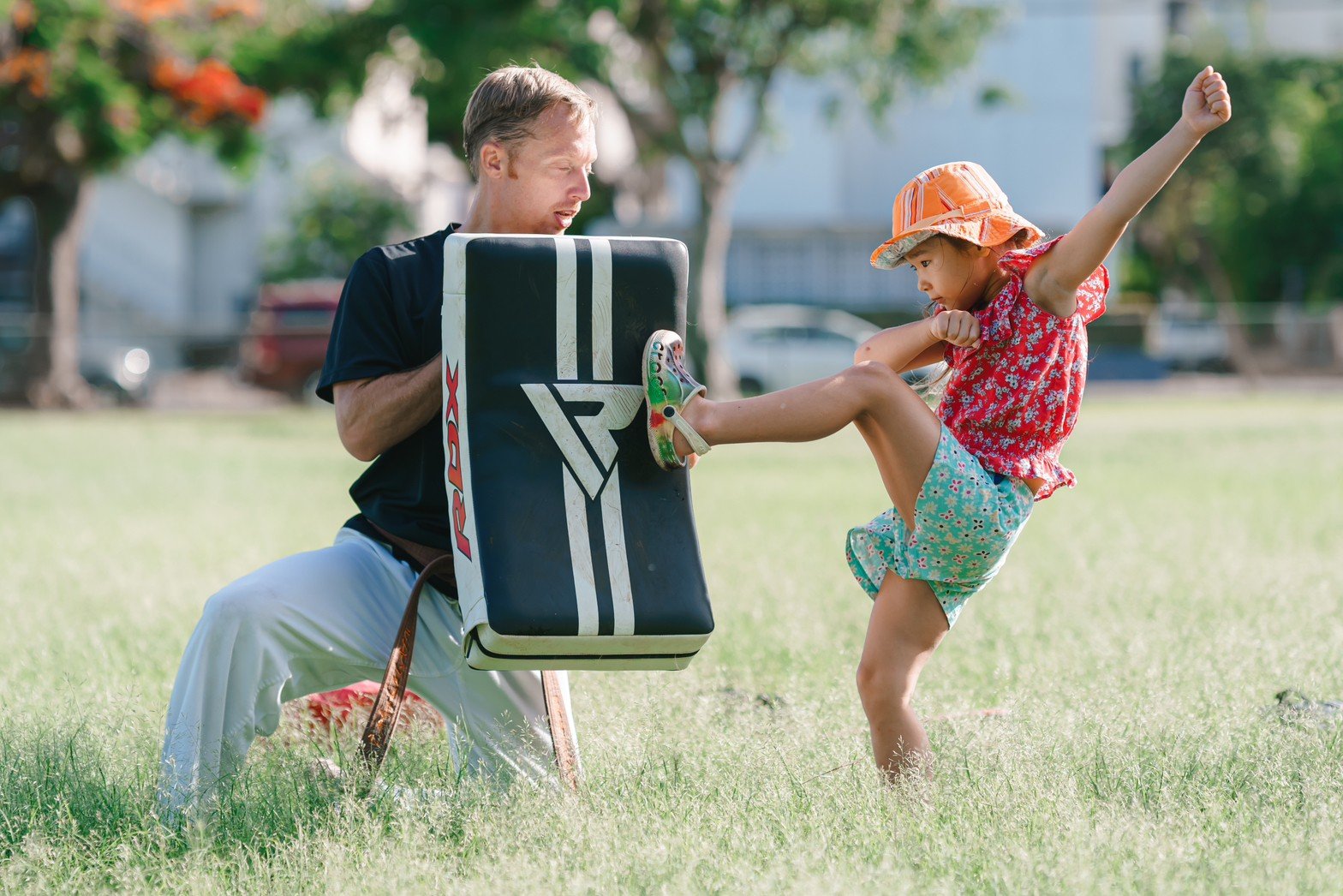 A young girl practicing a high kick toward a martial arts pad held by a man outdoors in a grassy area.