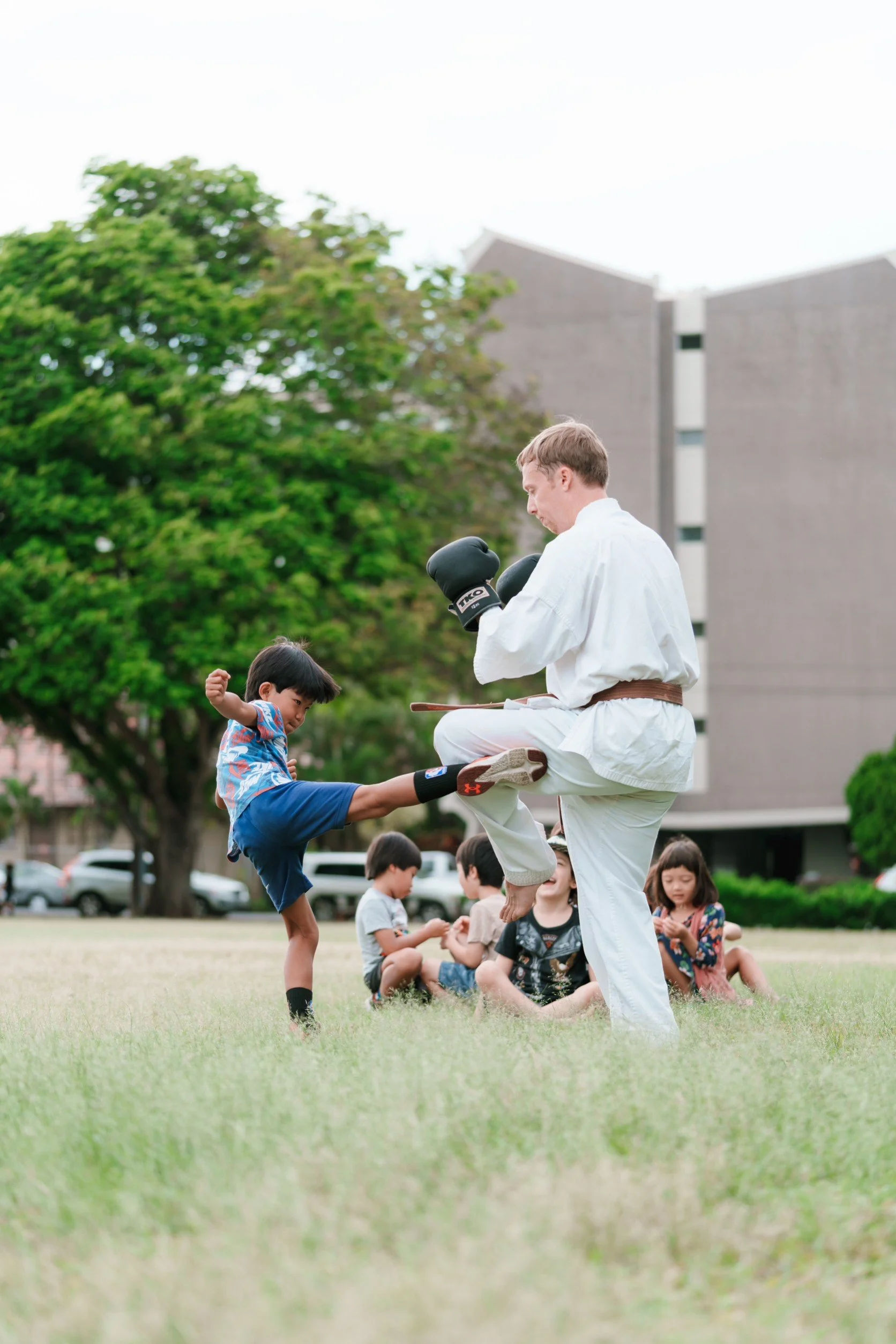 A martial arts instructor practicing martial arts kicks with a young boy while other children watch in a park.