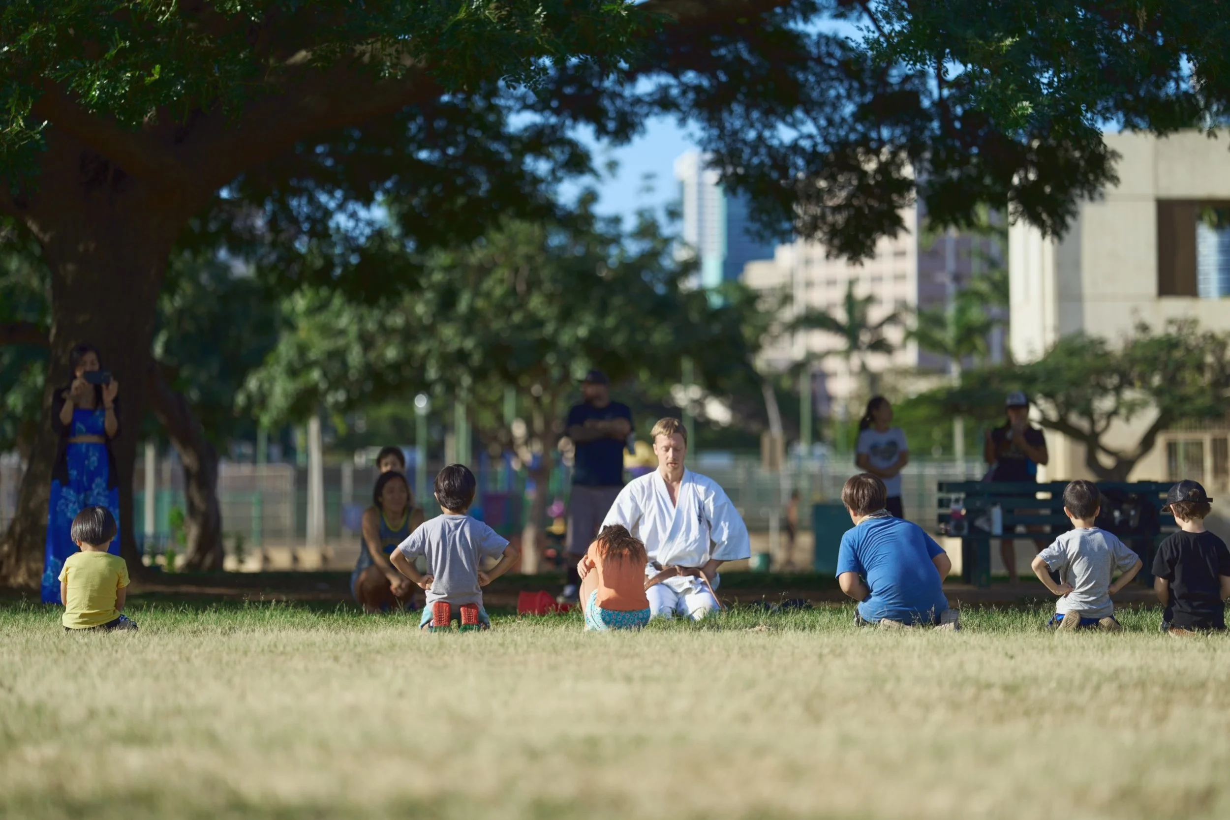 A martial arts class in a park, with children sitting on the grass in front of an instructor dressed in a white gi, under large trees with a cityscape in the background.