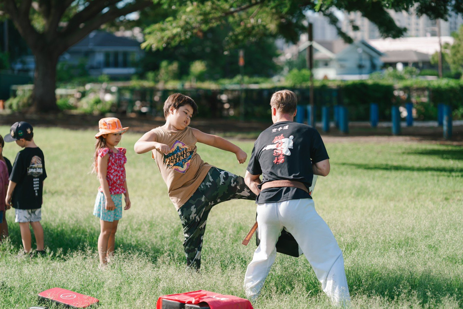 A martial arts instructor demonstrates a kick to a young girl practicing in a park on a sunny day. Other children are watching, slight shadows cast by the trees.
