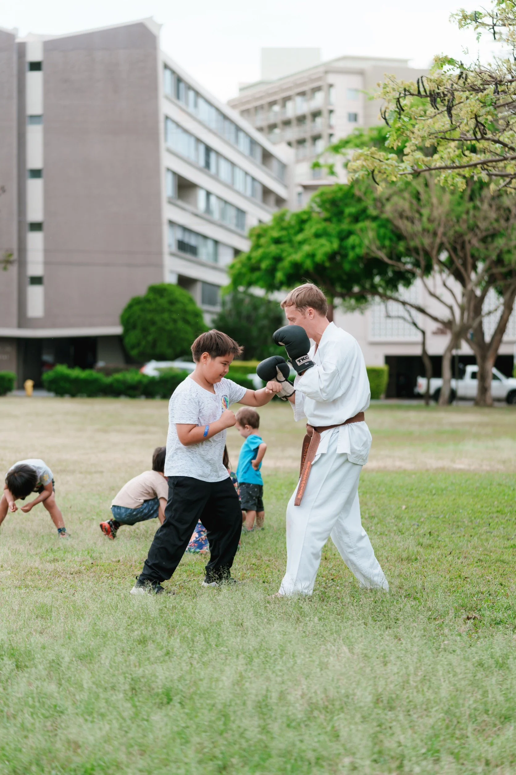 A man and a boy sparring in a martial arts practice, with other children in the background in an outdoor grassy area surrounded by apartment buildings and trees.