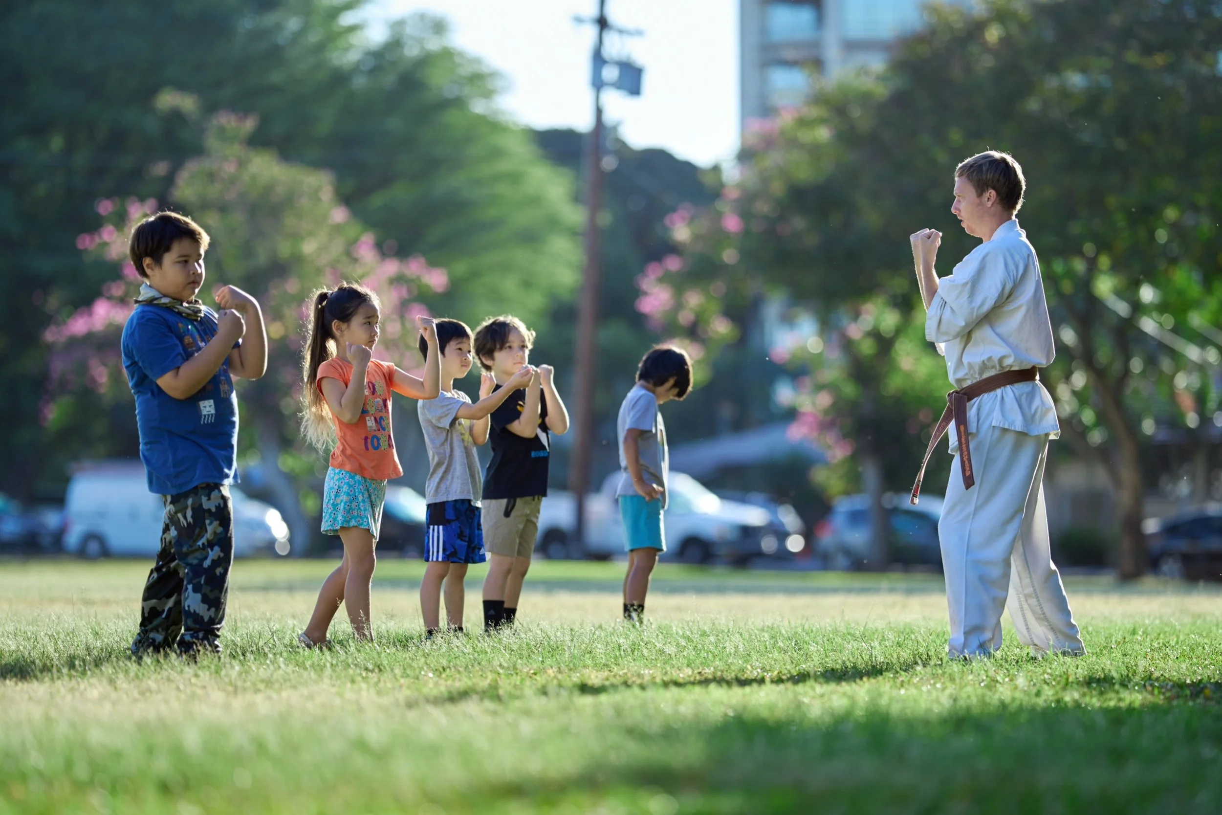 An adult in a martial arts uniform instructs children in a martial arts class outdoors on a grassy field with trees and cars in the background.