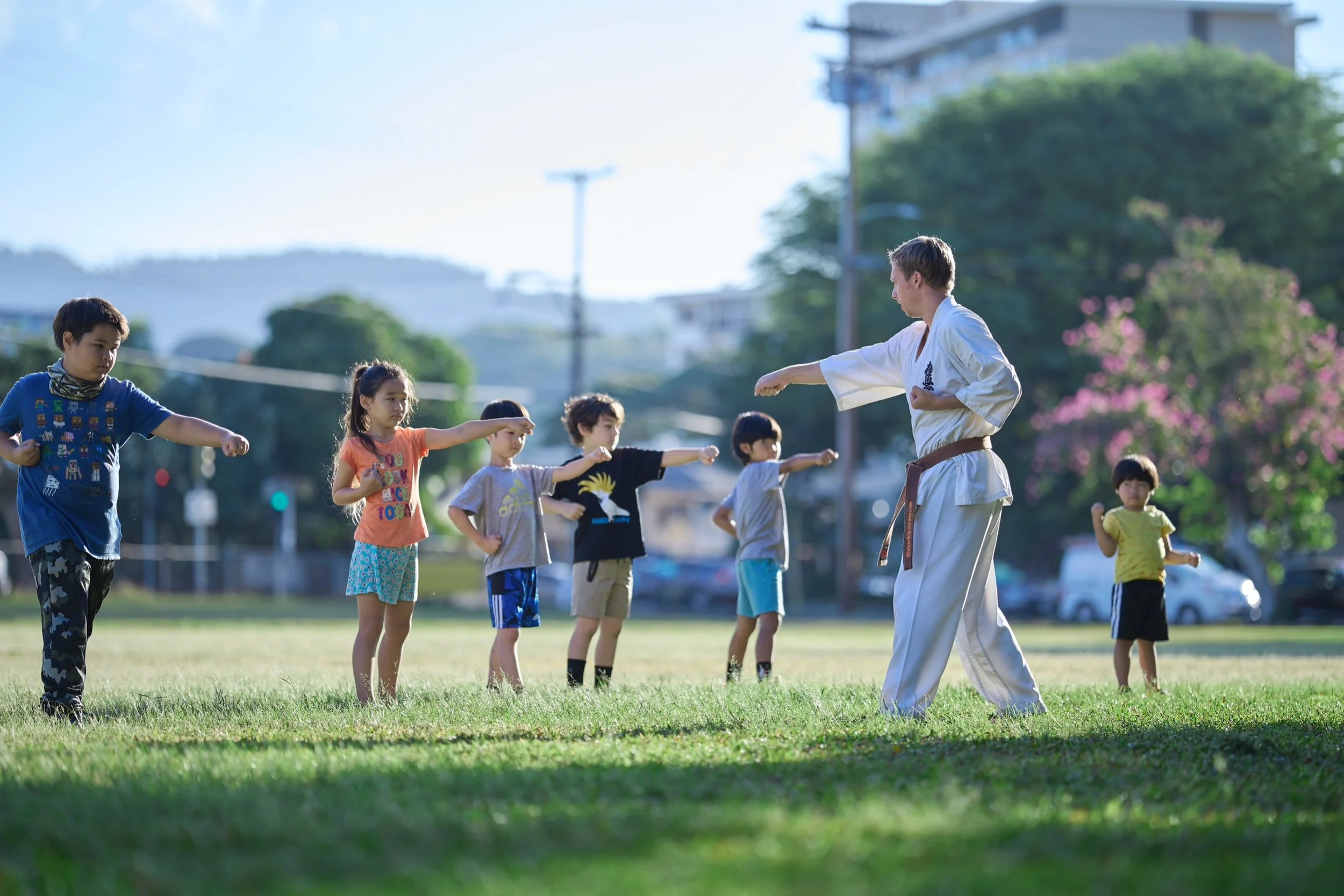 A martial arts instructor demonstrating a stance to young children outdoors on a grassy field during daylight.