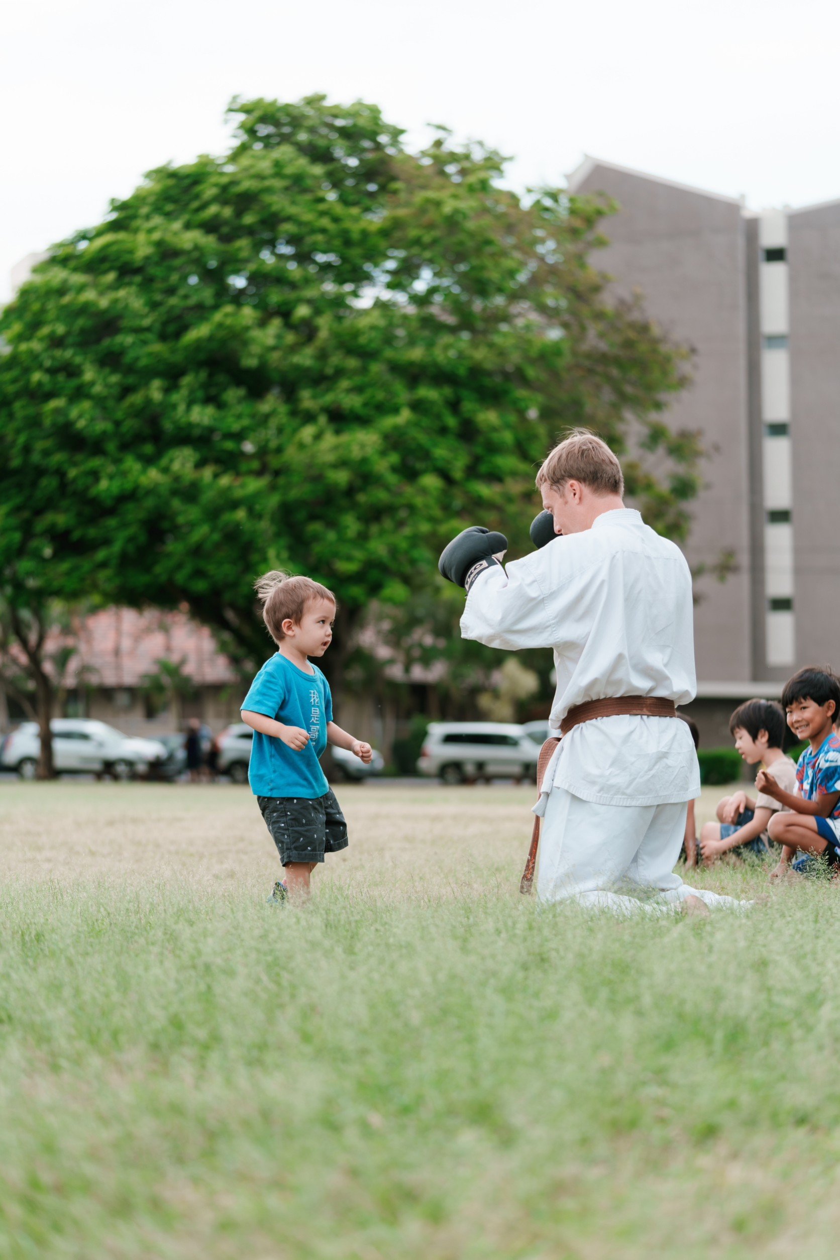 A man practicing karate with children in a park, a young boy in a blue shirt and black shorts facing him, and other children sitting on the grass watching.