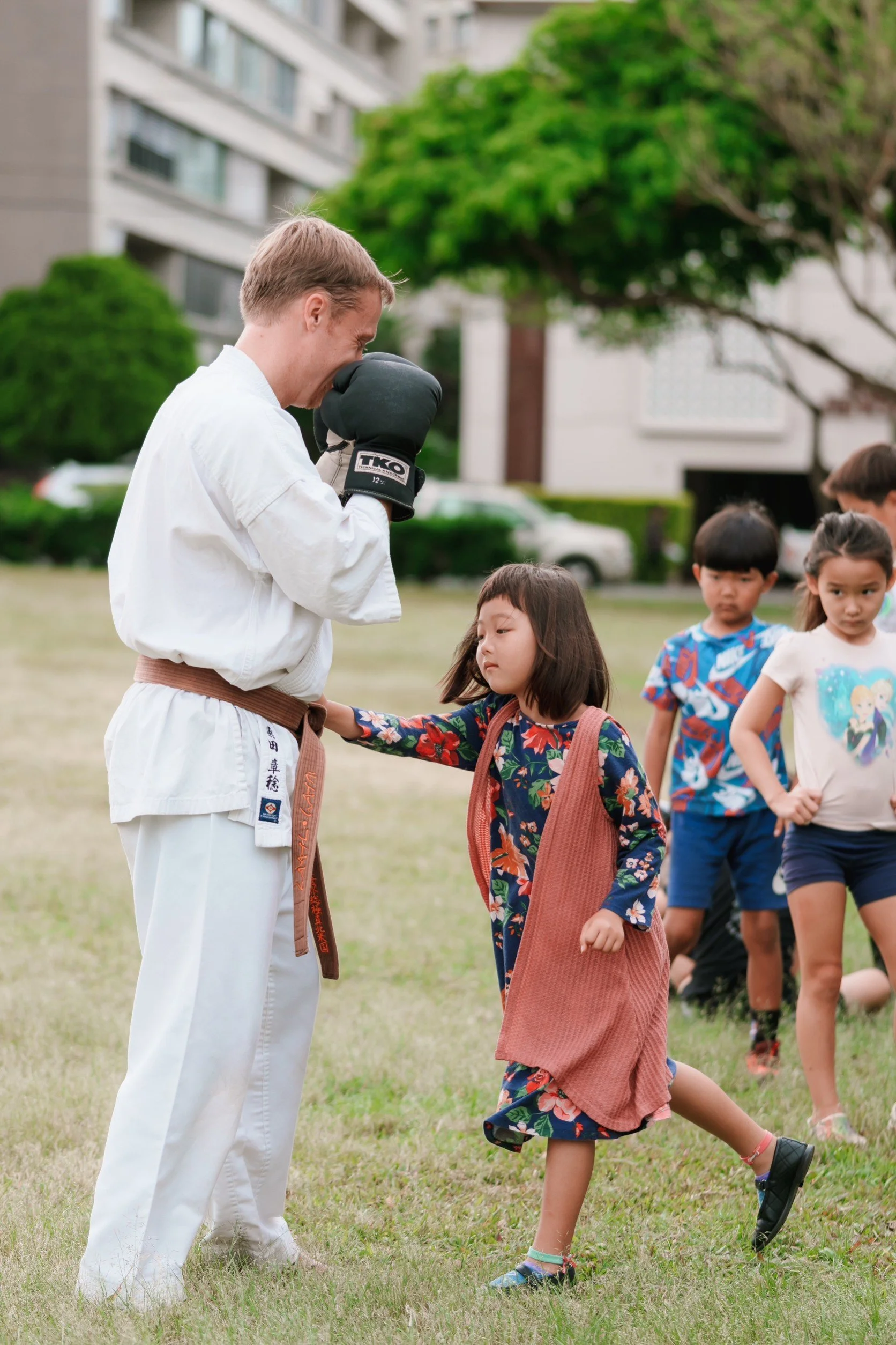 A martial arts instructor in a white uniform with a brown belt shows a young girl in a floral dress and pink vest how to block a punch during a class outside. Other children stand in line behind them, watching.