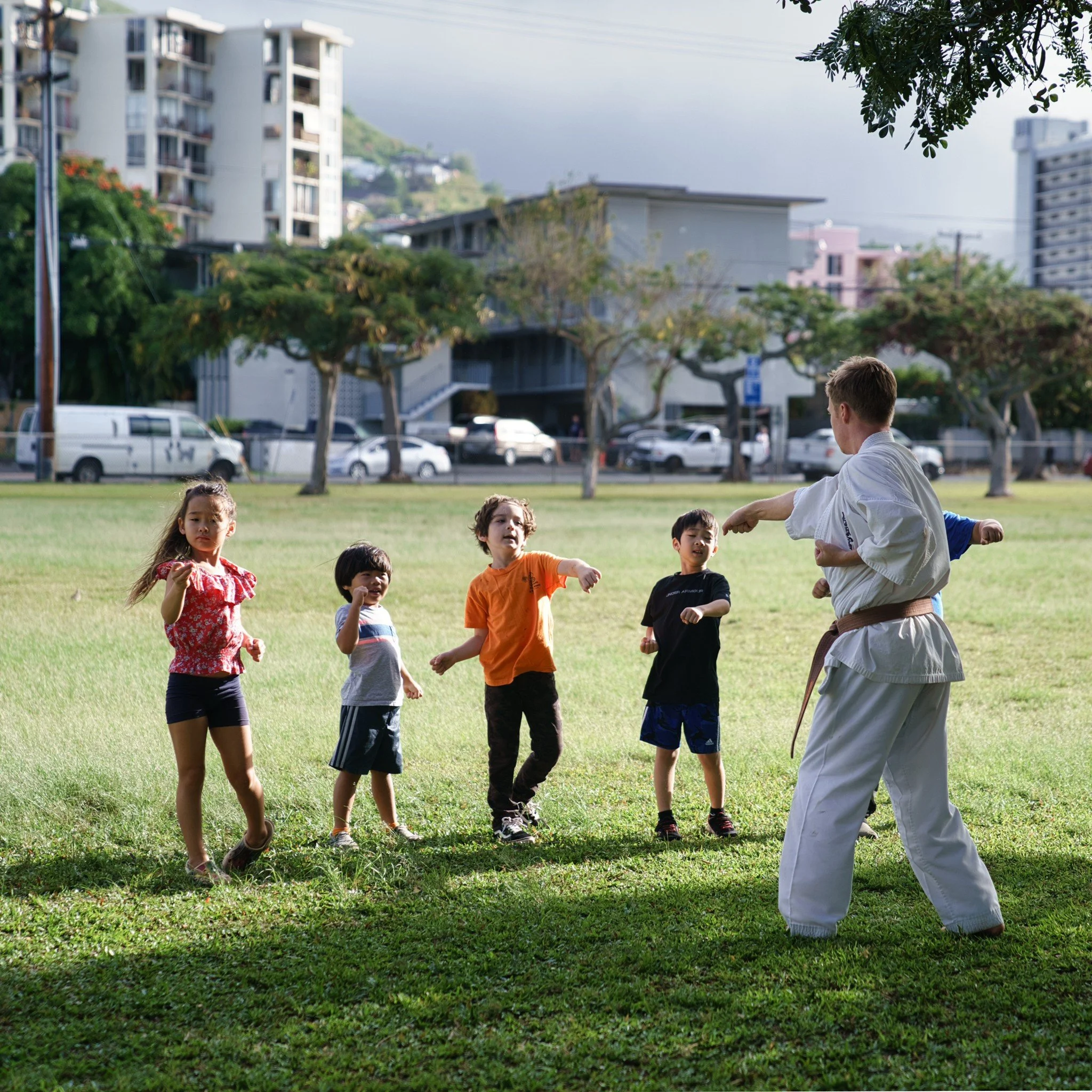 Children practicing martial arts in a park with a martial arts instructor in traditional uniform, surrounded by trees and urban buildings.