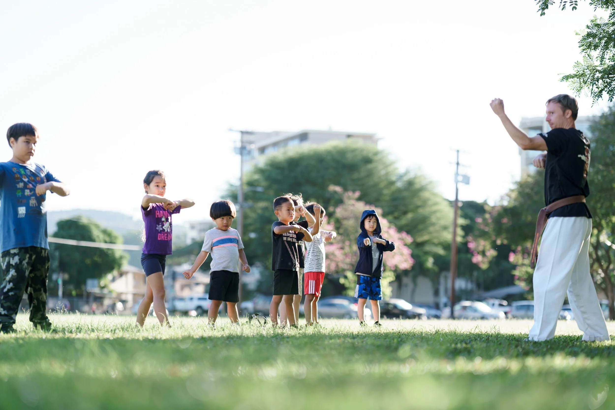 Group of children practicing martial arts outdoors while an instructor demonstrates. The children are standing on grass with trees and buildings in background.