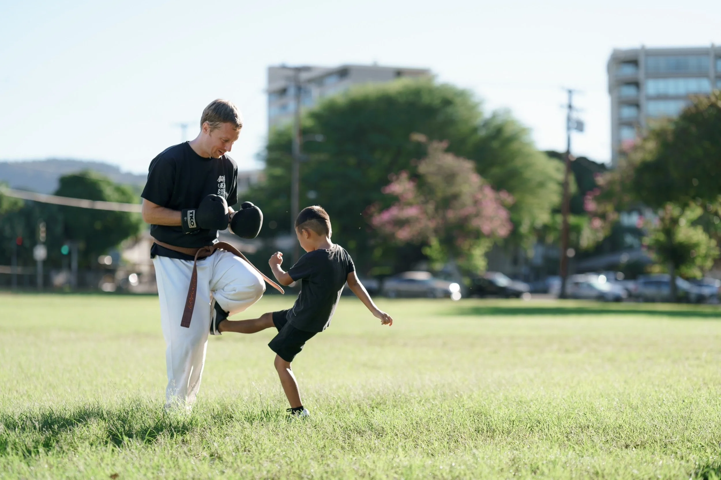 A man in traditional martial arts uniform practicing martial arts with a young boy outdoors on a grassy field in a park, with trees and buildings in the background.