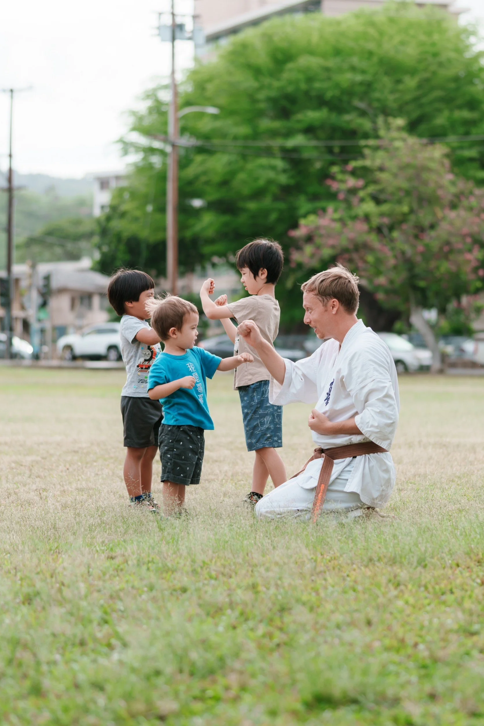 A martial arts instructor in a white gi and brown belt demonstrates a move to four young children in a park on a grassy field with trees and buildings in the background.