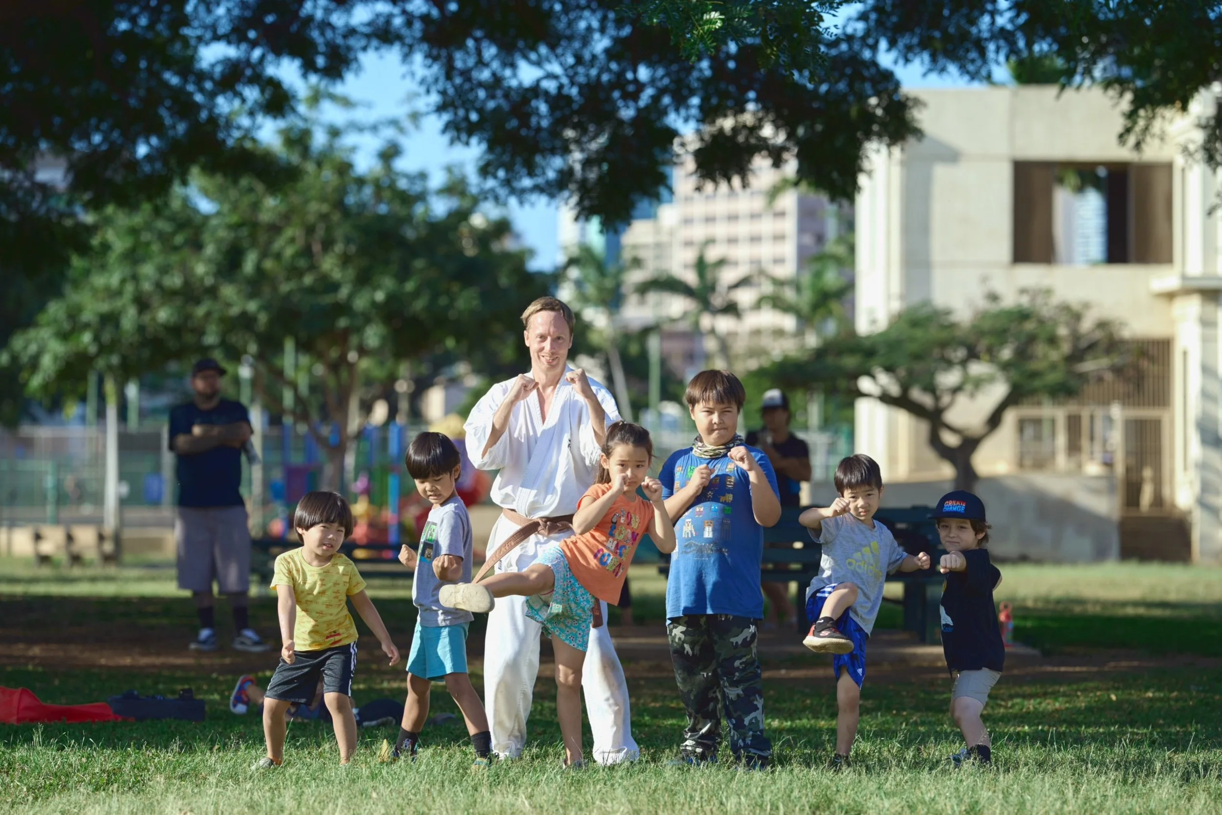 A martial arts instructor and six children practicing martial arts moves outdoors in a park, with trees and buildings in the background.