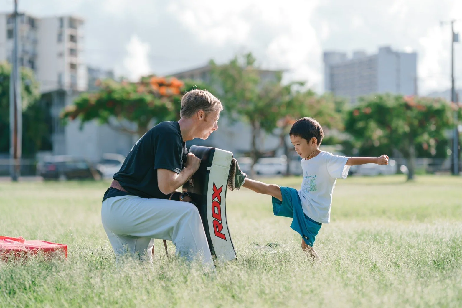 An older man practicing martial arts with a young boy outdoors in a park on a sunny day.