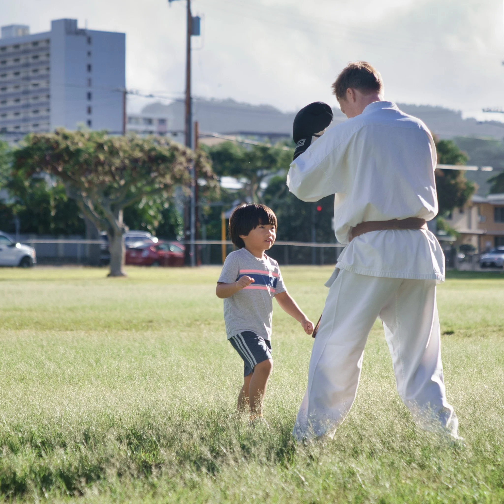 A young boy learning martial arts from an instructor outdoors on a grassy field in a city park.