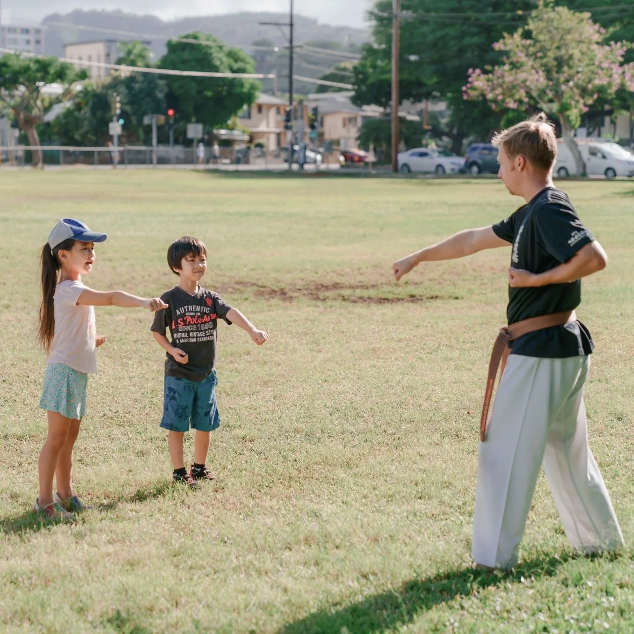A man in a martial arts uniform demonstrating a punch move to two children in a park.