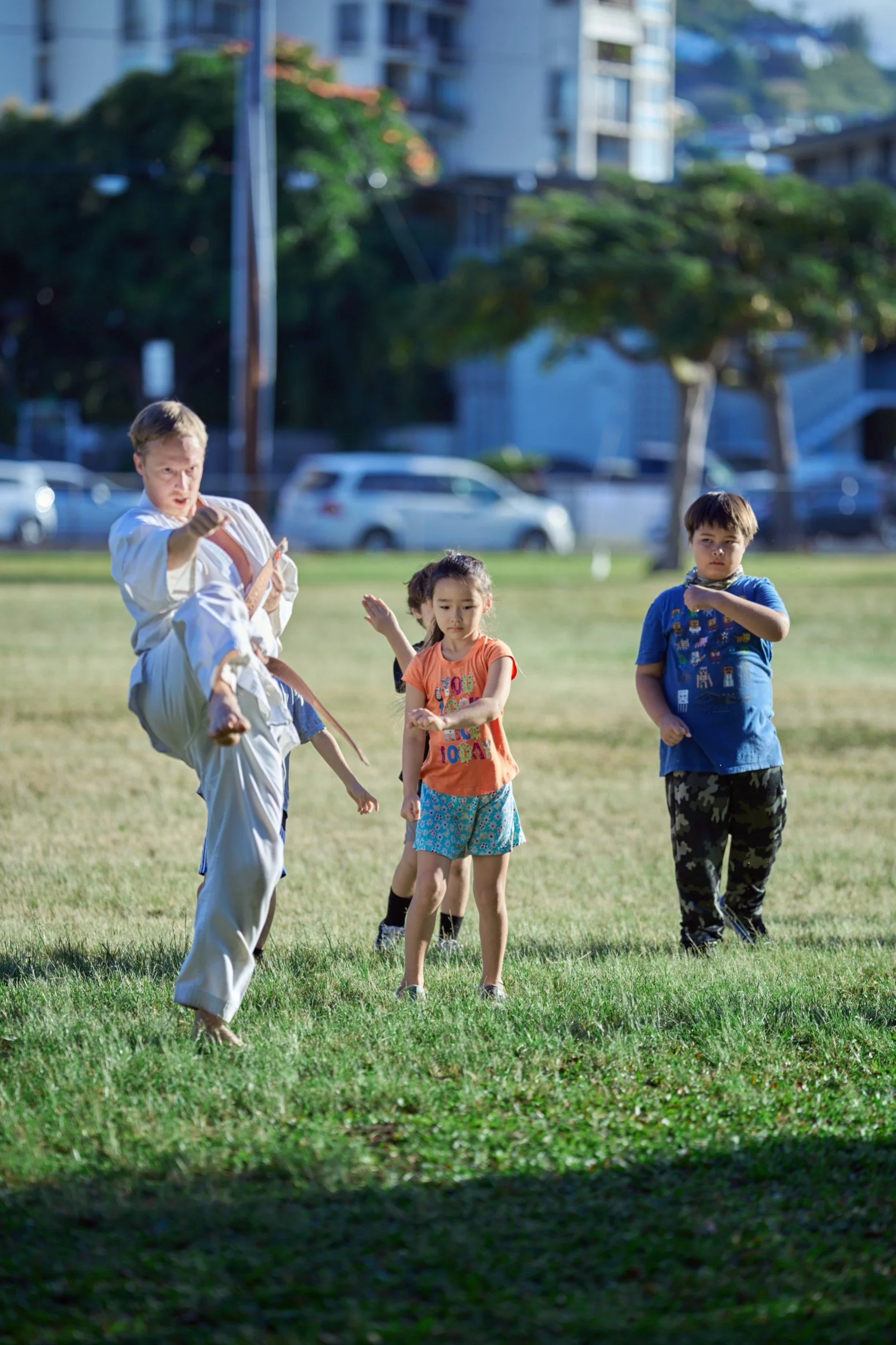A martial arts instructor demonstrating a kick to a group of children in a park on a sunny day.