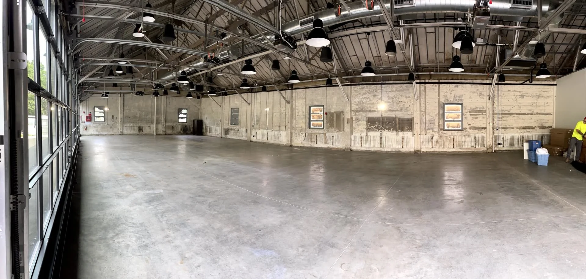 Empty industrial-style space with high ceilings, exposed metal beams, and black hanging industrial lights. The concrete floor is polished, and there are several small windows along the wall. A person in a yellow shirt is standing near a stack of blue containers on the right side.