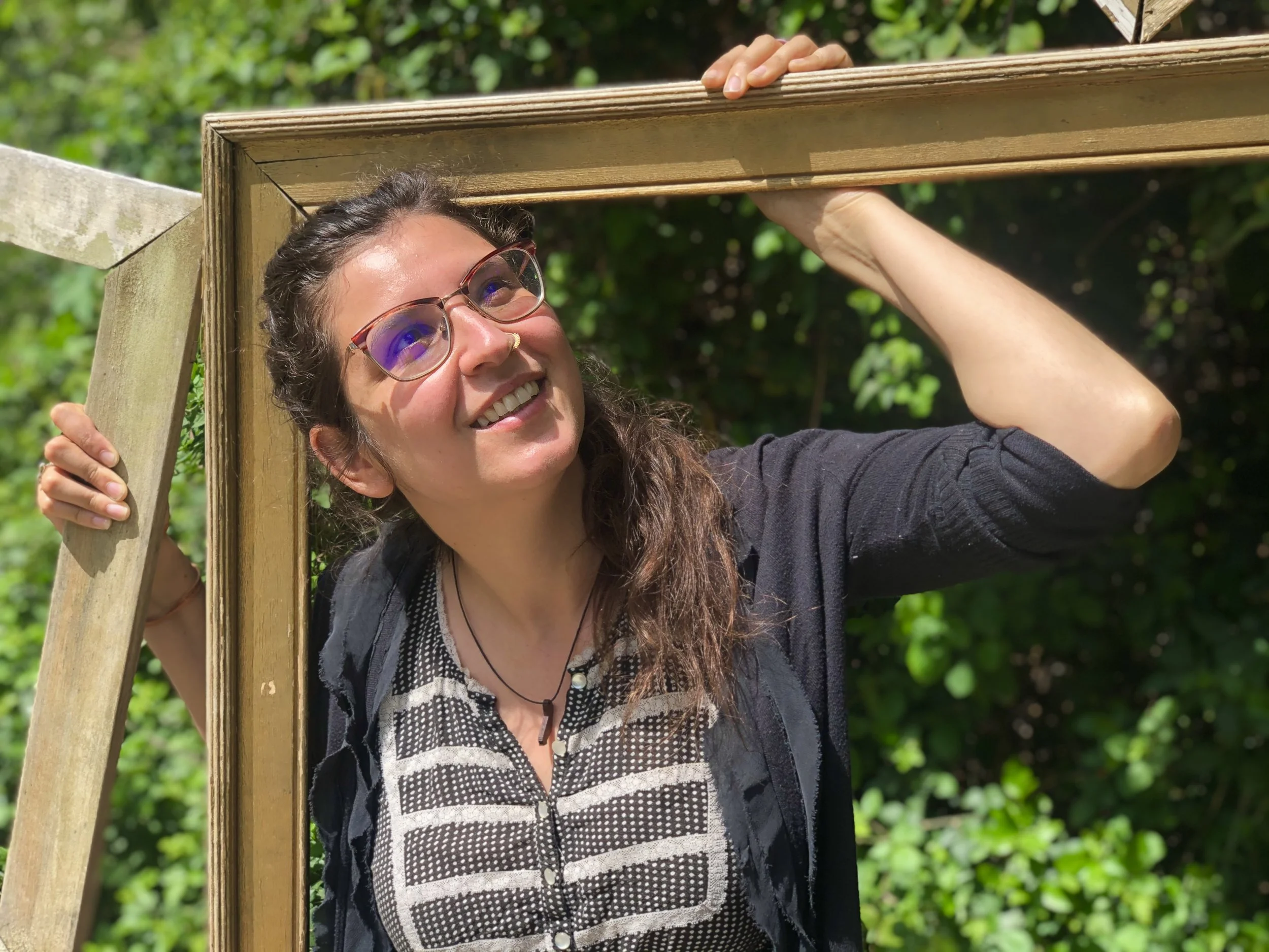 A woman with glasses, long dark hair, and a nose ring smiling outdoors while holding a wooden frame.