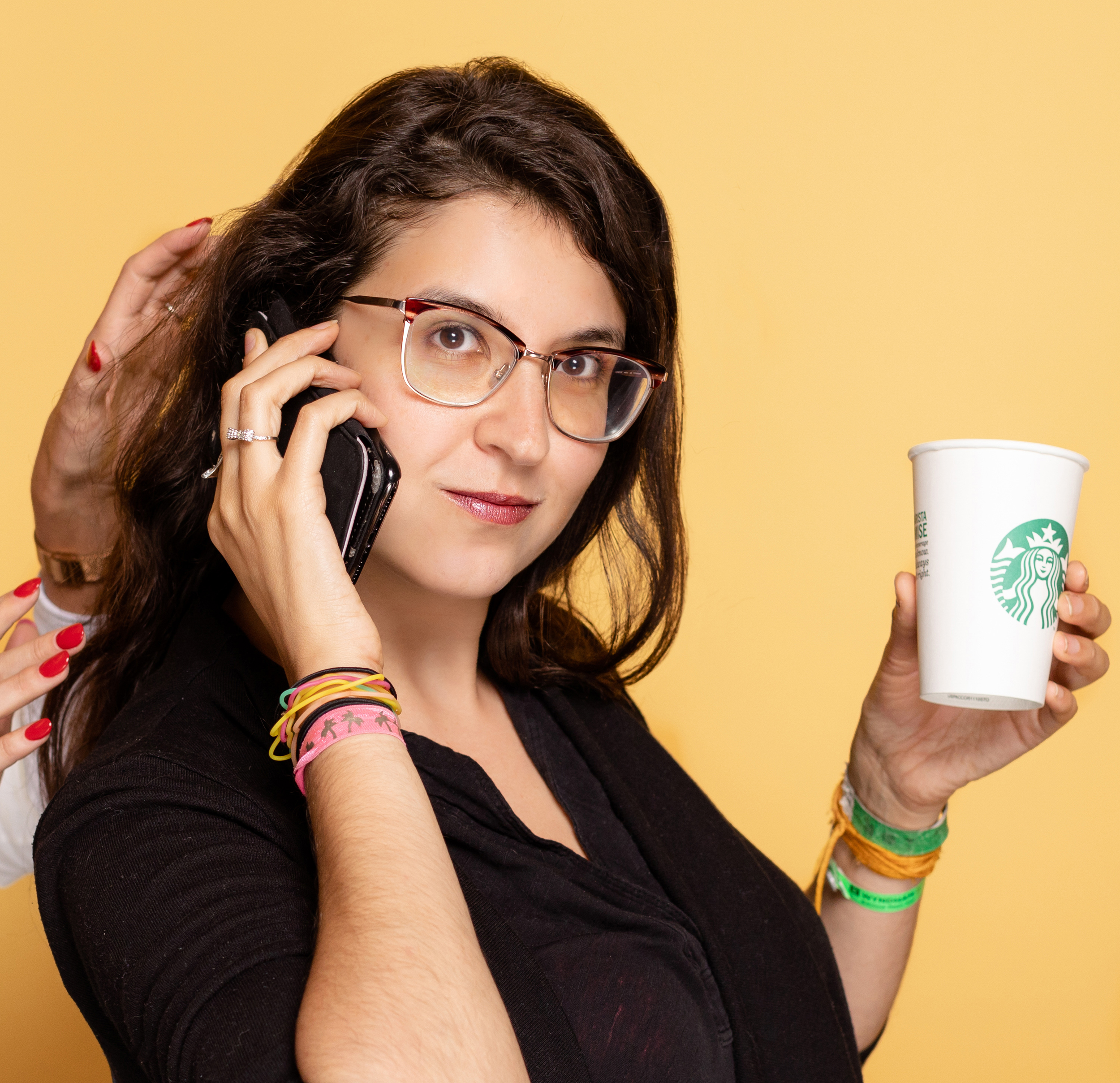 A woman with brown hair and glasses holding a Starbucks cup in one hand and talking on a cellphone with the other.