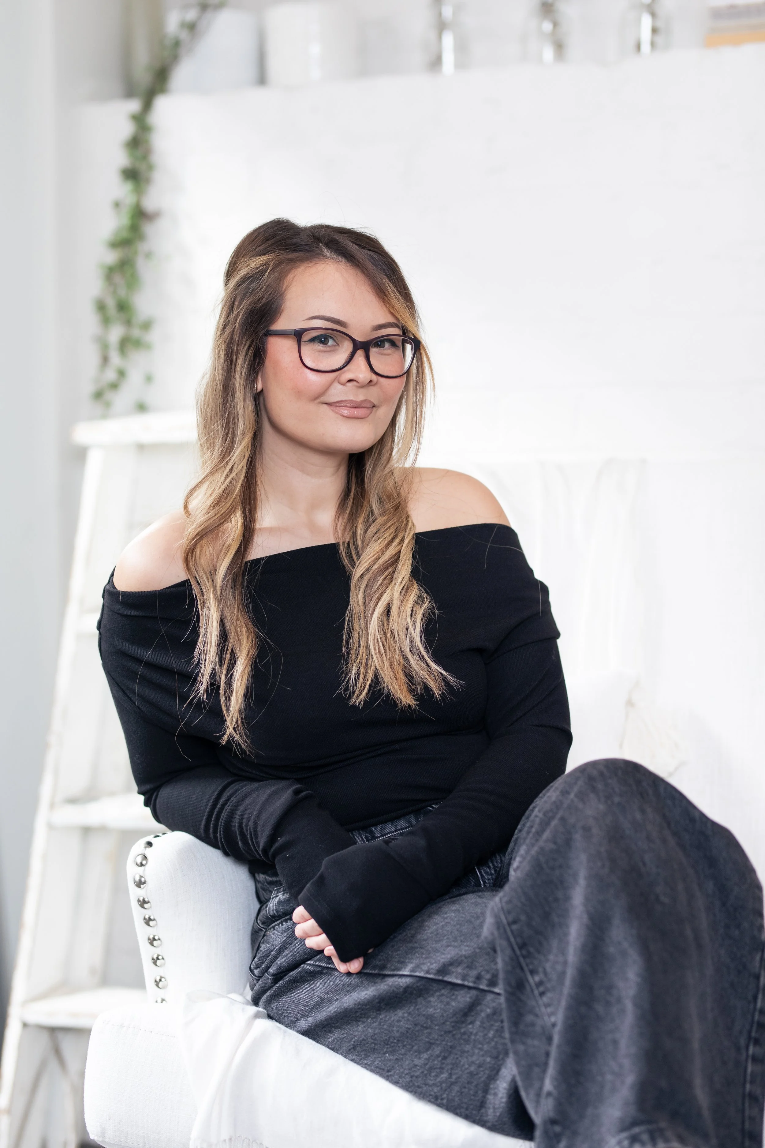 A young woman with glasses and long, wavy hair sitting on a white chair in a bright room with white walls and a ladder in the background, looking at the camera with a neutral expression. Makeup artist wellington NZ. Flawless soft glam makeup look.