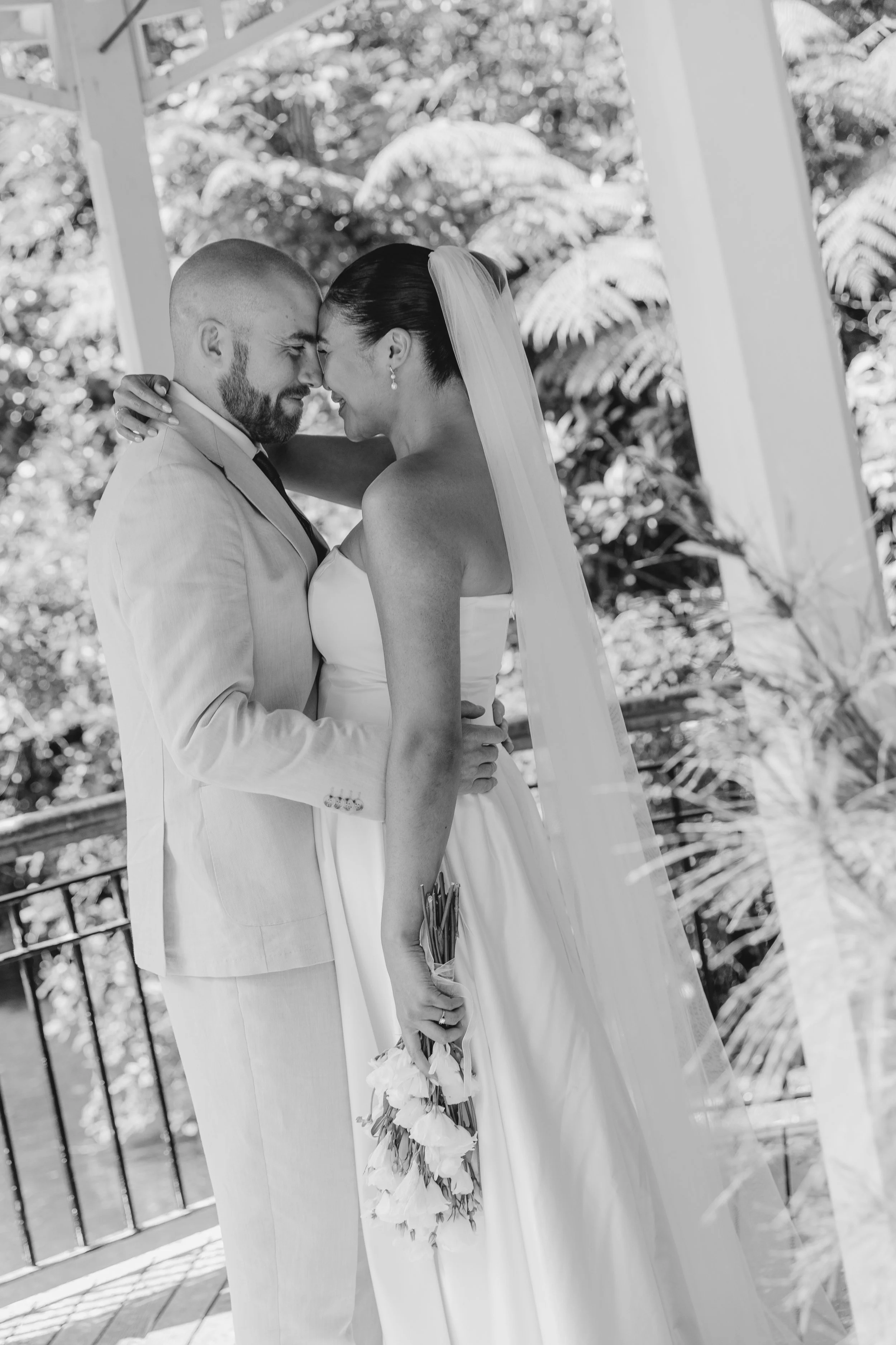 Black and white photo of a bride and groom standing close, touching foreheads and smiling, outdoors surrounded by trees and a metal fence. Bridal makeup for weddings. Natural glam, soft glam, beautiful. In wellington new zealand