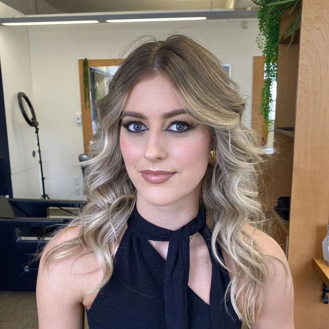 Young woman with blonde, curly hair and striking blue eyes, wearing makeup and a black sleeveless top with a bow tie around her neck, posing indoors in a well-lit room with wooden furniture and green plants. Full glam makeup. Bridal makeup. beautiful