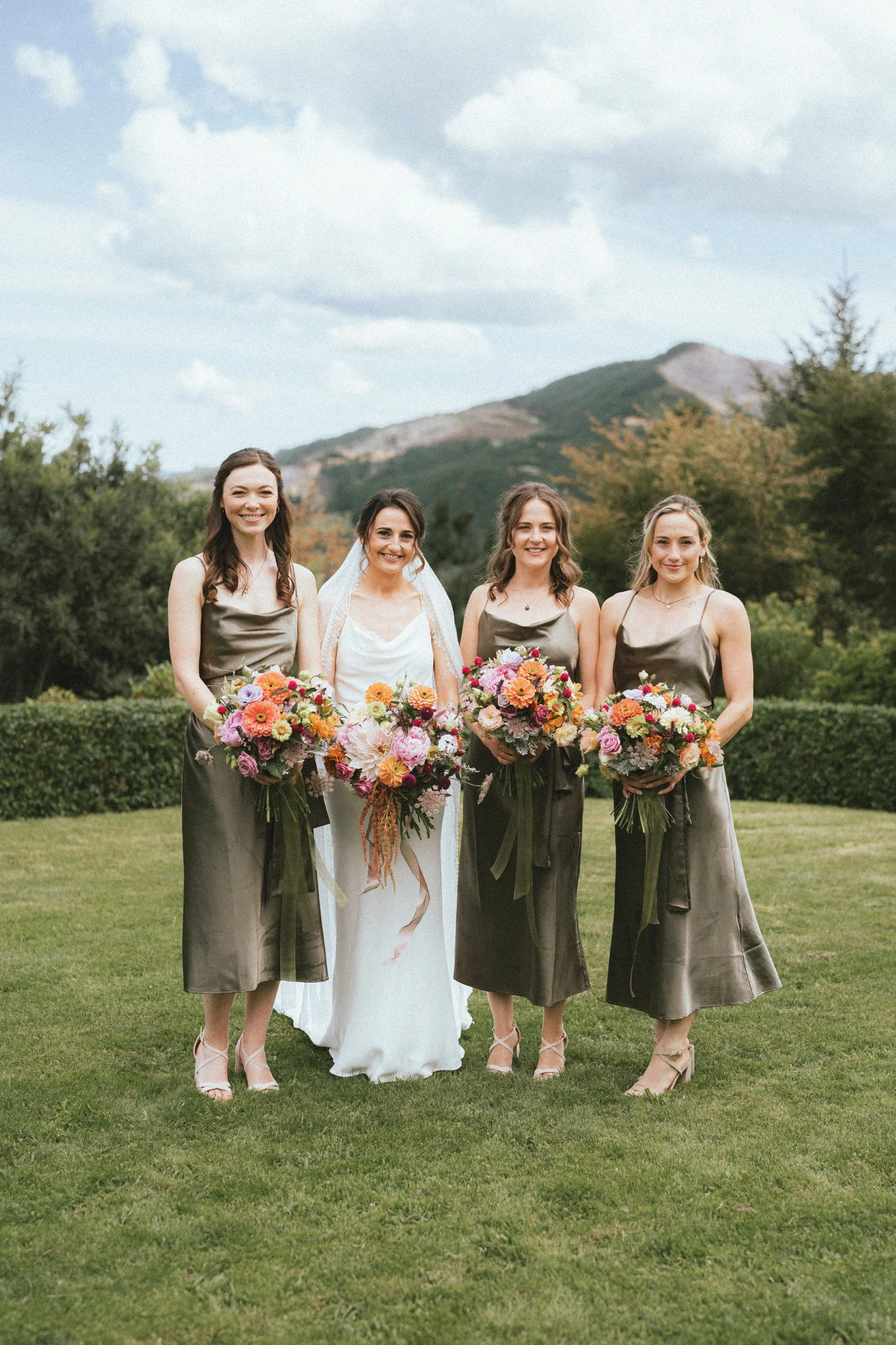 A bride and three bridesmaids standing outdoors on a grassy field with mountains and trees in the background, all holding large colorful floral bouquets and dressed in formal attire. Soft glam natural makeup look. No makeup makeup look