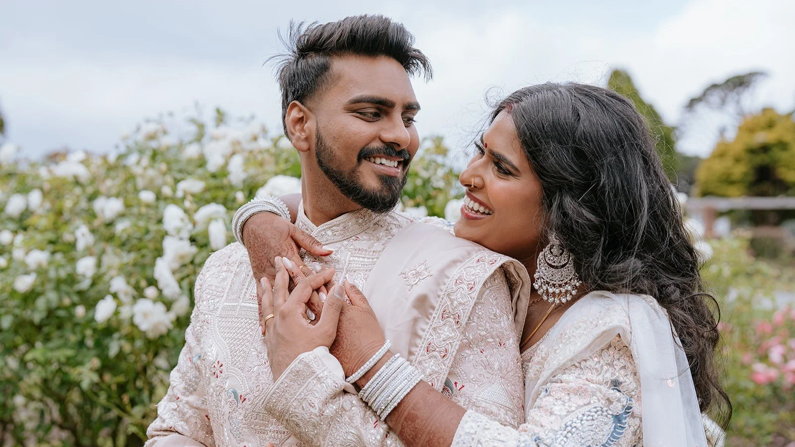 A joyful Indian couple in traditional wedding attire embracing in a garden with white flowers, smiling at each other. Indian wedding. Makeup was done by makeup by lucy loo on the Indian bride. Asian bride flawless makeup. Wellington New Zealand