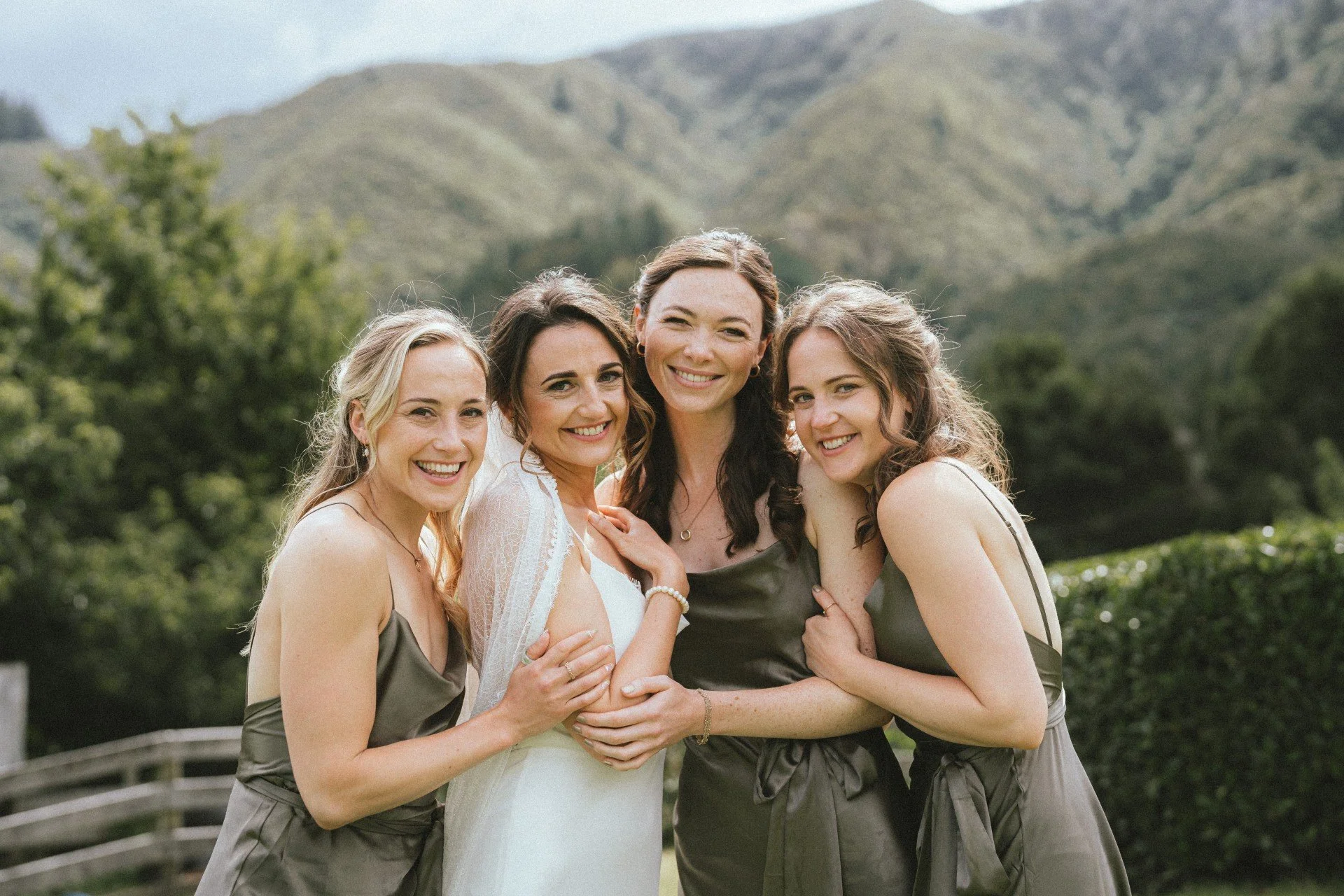A group of five women smiling and embracing each other outdoors with mountains and trees in the background, dressed for a wedding or formal occasion. bride with 3 bridesmaids. Stunning flawless bridal makeup. Makeup for any event or special occasion