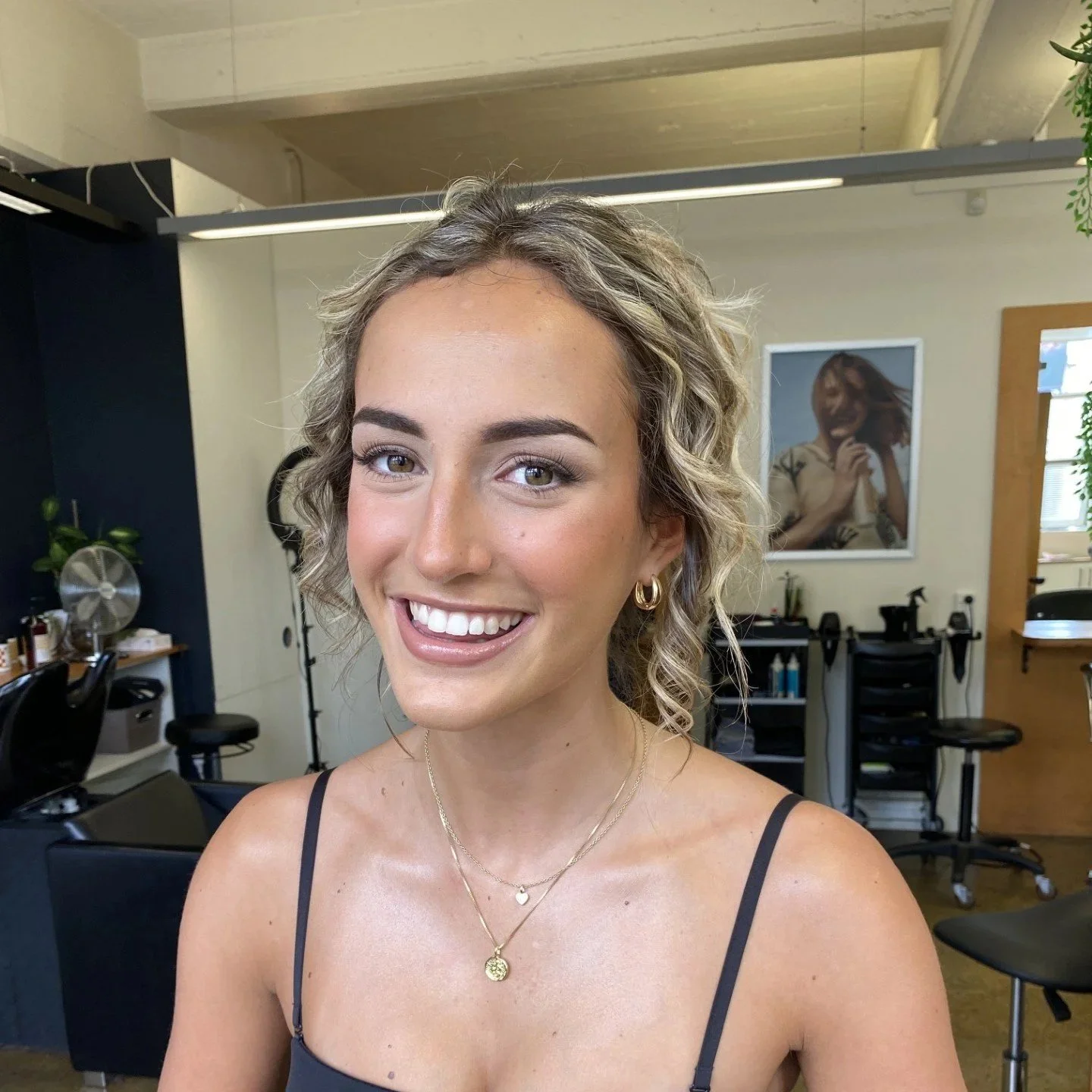 A woman with long wavy blonde hair wearing natural makeup and a black sleeveless top, standing in a hair salon. natural glam makeup. Bridal makeup. beautiful natural makeup look