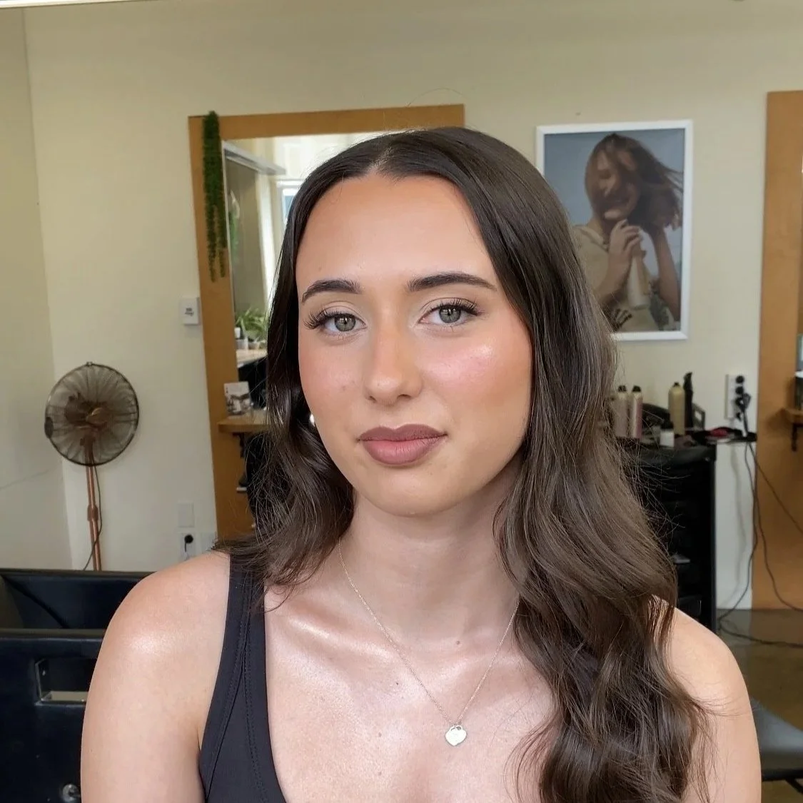 A woman with long wavy brown hair wearing natural makeup and a black sleeveless top, standing in a hair salon. soft glam makeup. Bridal makeup. beautiful natural makeup look