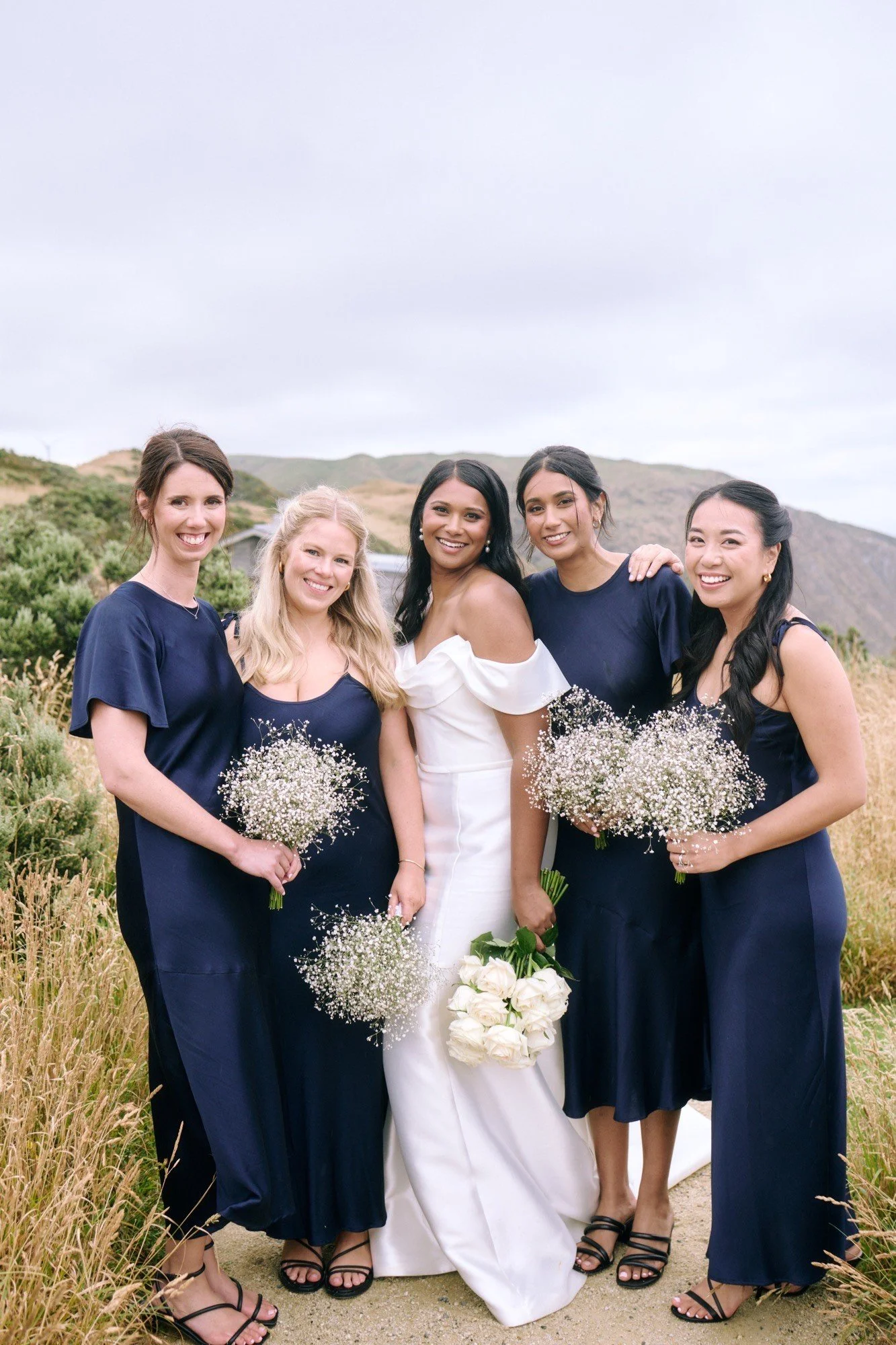 A group of five women, including a bride in a white wedding dress holding a bouquet of white roses, standing outdoors on a grassy field with hills in the background. Four women around her are dressed in navy blue dresses, holding small bouquets of wh