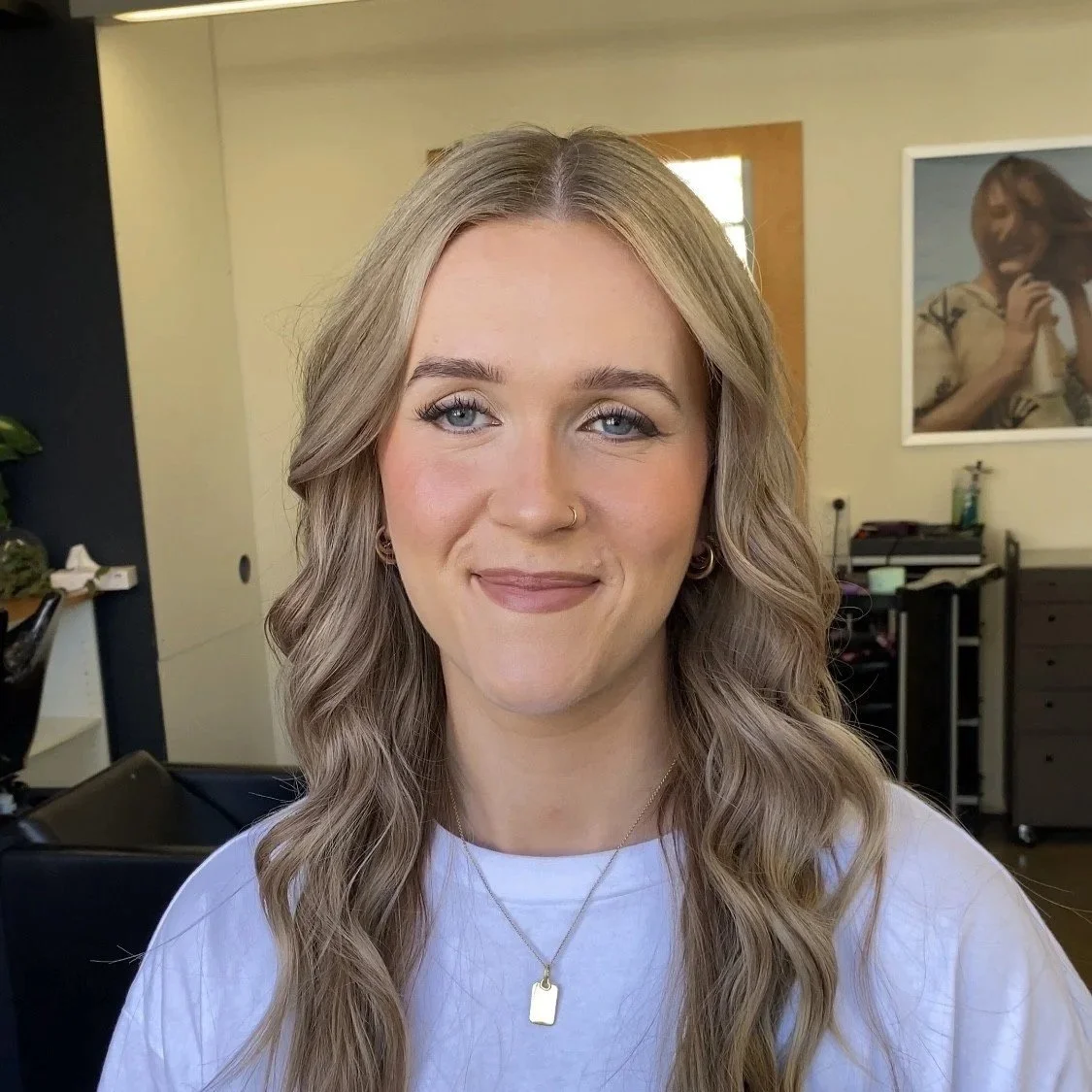 A young woman with long wavy blonde hair, wearing a white shirt and gold jewelry, smiling in a hair salon. soft glam makeup. Bridal makeup. beautiful natural makeup look. bridal specialist. Wedding makeup
